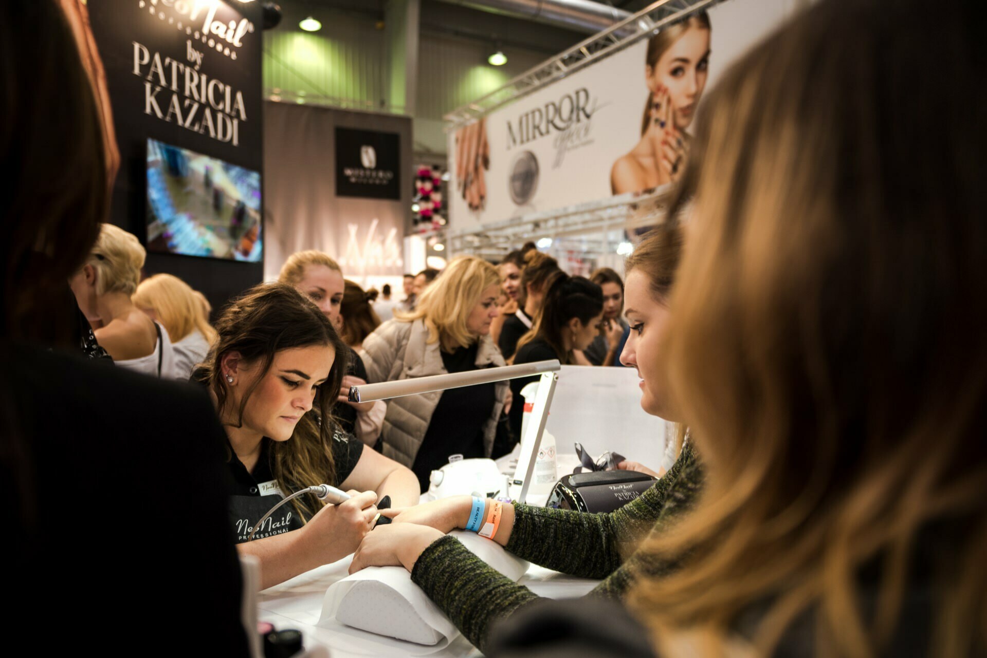 A crowded beauty exhibition with a beautician giving a manicure to a seated client, captured by Marcin Krokowski, a Warsaw-based event photographer. Several people in the background are observing and engaging with various booths and promotional displays. Prominent signs and advertisements are visible.  