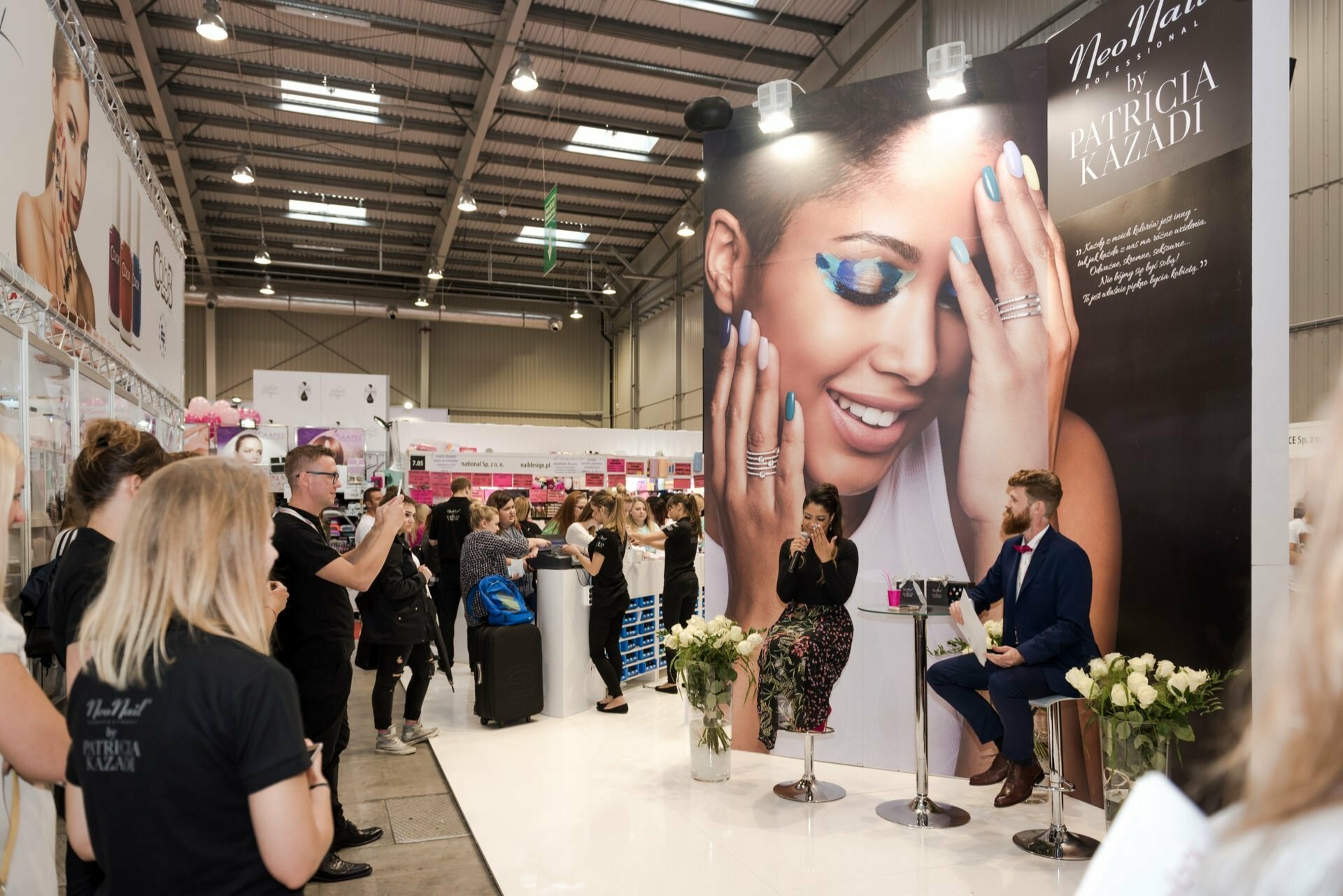 At the crowded indoor event, people gathered around a booth with a large poster depicting a woman in blue makeup, promoting Patricia Kazadi's product. Marcin Krokowski, an acclaimed event photographer, captures a scene in which two people speak into a microphone while spectators take pictures. 