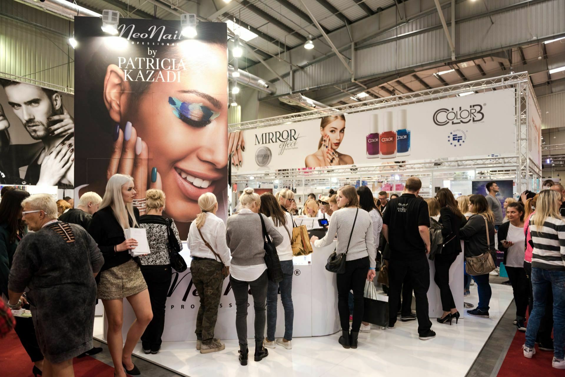 At a cosmetics fair, you can see a large gathering of people with booths advertising various cosmetic products. A prominent banner shows a smiling woman with blue eye makeup for NeoNail by Patricia Kazadi. Marcin Krokowski, a well-known Warsaw event photographer, captures attendees as they interact with exhibitors and browse the display.  