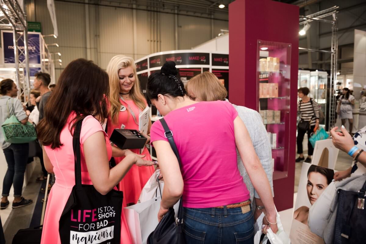 Grupa kobiet gromadzi się wokół stoiska na konwencji lub targach i wydaje się zainteresowana produktem prezentowanym przez kobietę w różowej sukience. Dobrze oświetlone i ruchliwe stoisko udekorowane jest produktami kosmetycznymi i pielęgnacyjnymi, a wszystkie zdjęcia wykonał Marcin Krokowski, doświadczony fotograf w Warszawie.