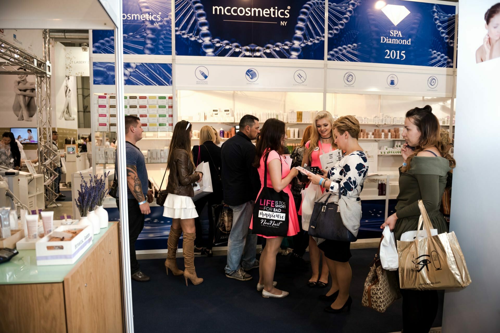 People gather around a booth displaying skin care products and cosmetics at the fair. At the booth, labeled "MCCosmetics NYC," various products sit on shelves and promotional signage is displayed above. Attendees discuss and look at the products, while event photographer Marcin Krokowski captures fleeting moments.  