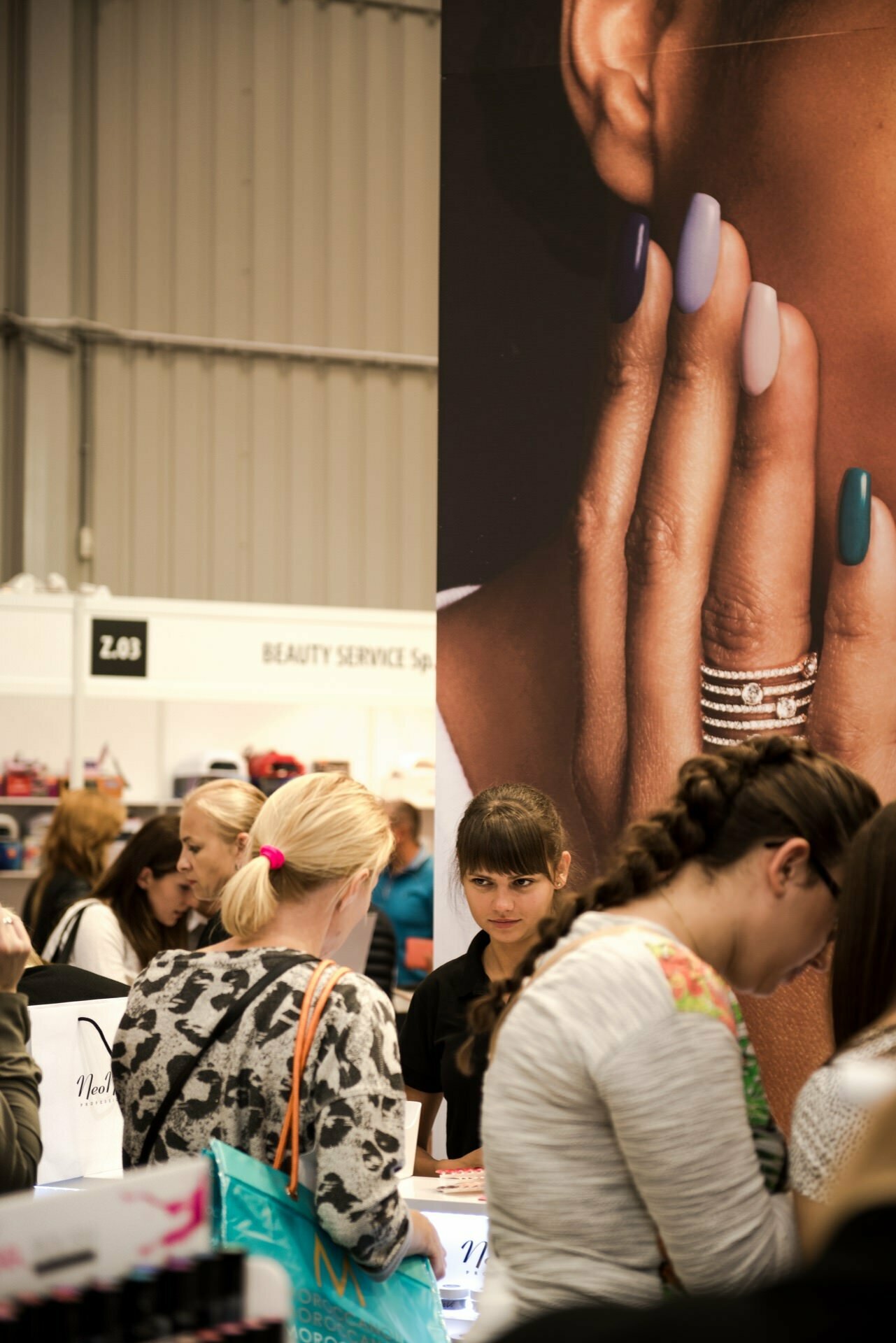 A crowded cosmetics fair where people are looking at various products. In the foreground, individuals look at the items on display, while event photographer Marcin Krokowski captures the scene. In the background, a large poster depicts manicured nails in various colors. The atmosphere is full of tension and focus.   