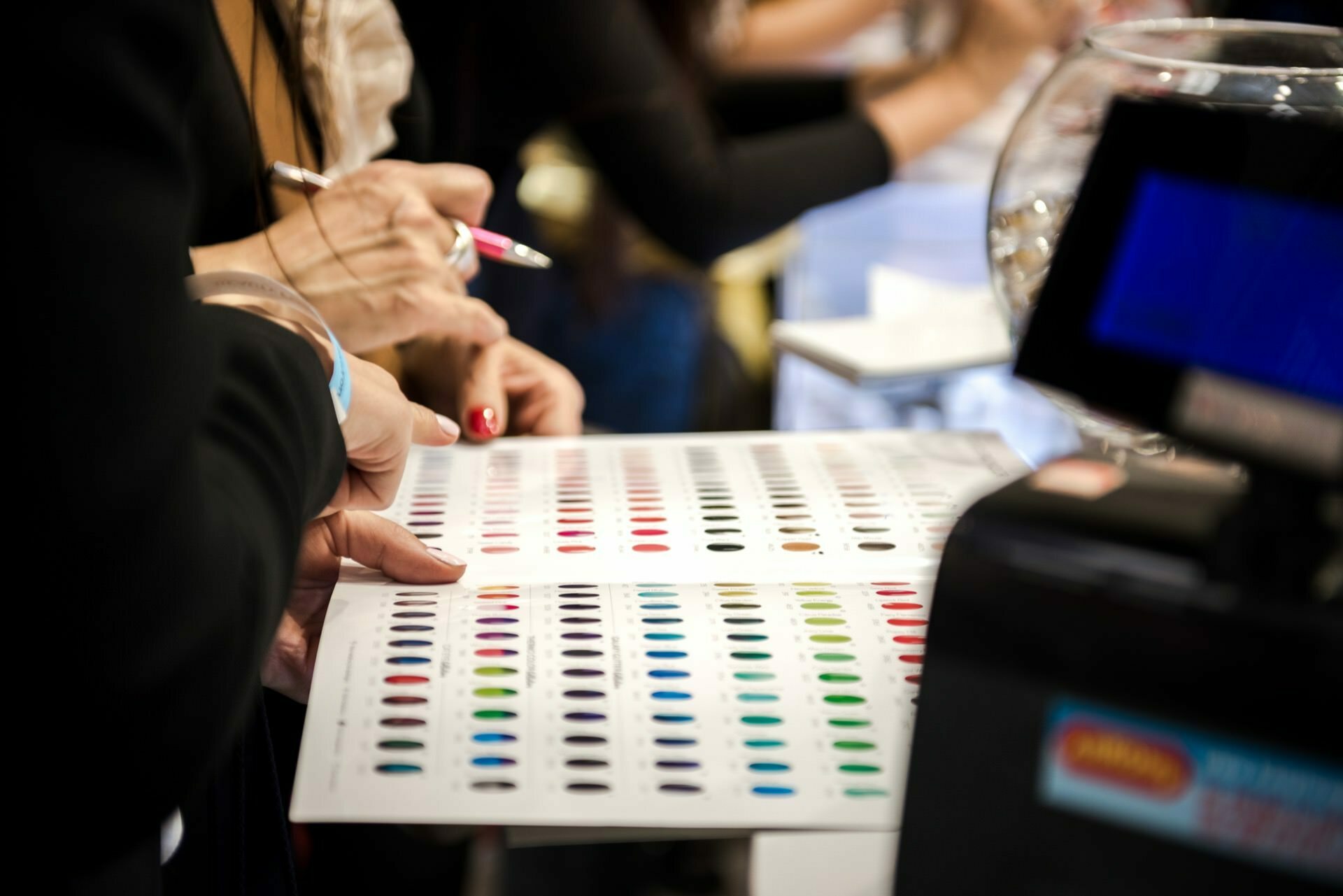 People reviewing a color selection chart on a stand containing numerous samples. One person is holding a pen and presumably taking notes or making selections. To the right of the photo, a digital screen is partially visible, capturing the meticulous work of acclaimed event photographer Marcin Krokowski.  