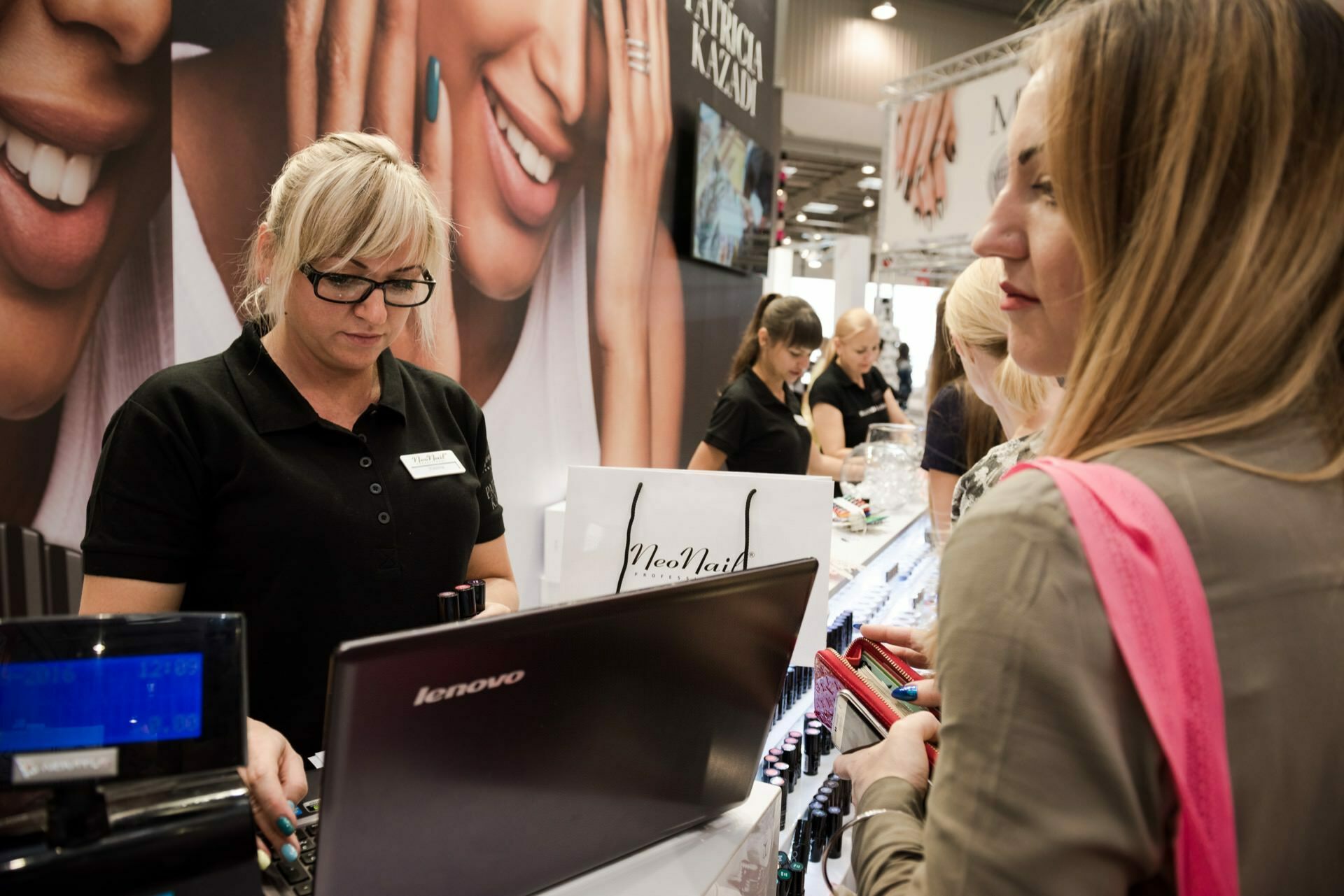 A woman wearing glasses and a black shirt serves customers at a beauty product stand. She stands behind the counter with a Lenovo laptop and bottles of nail polish. The background is decorated with posters showing hands with painted nails, beautifully captured by Marcin Krokowski, a photographer in Warsaw. Other booth employees work nearby.   