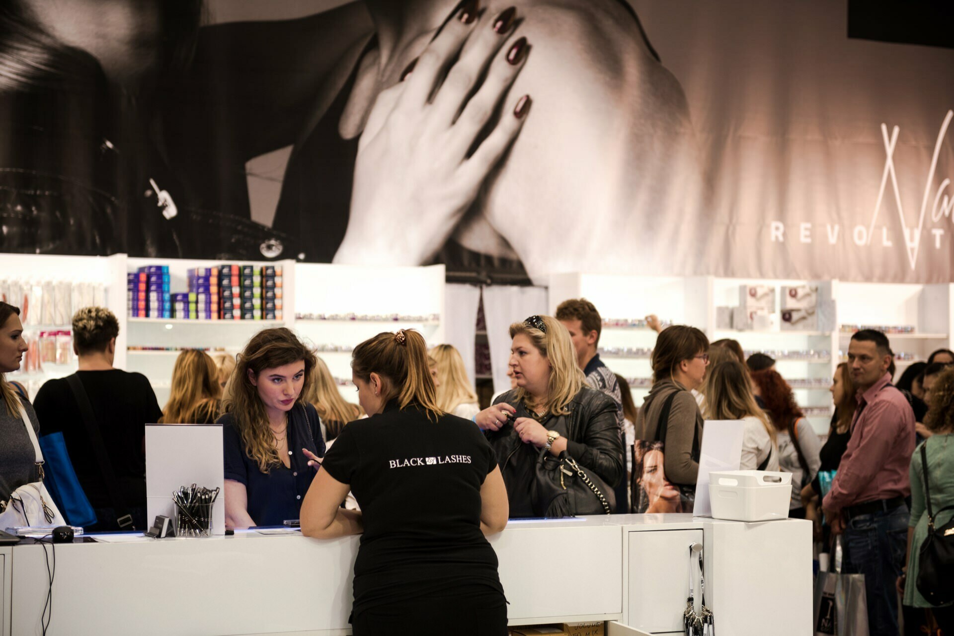 A group of people gather at the counter of a cosmetics store. A customer is being helped by a woman in a black shirt with the words "BLACK LASHES" written on it. In the background are shelves with various products, and on the wall is a large black and white promotional poster, beautifully captured by Marcin Krokowski, a photographer in Warsaw.  