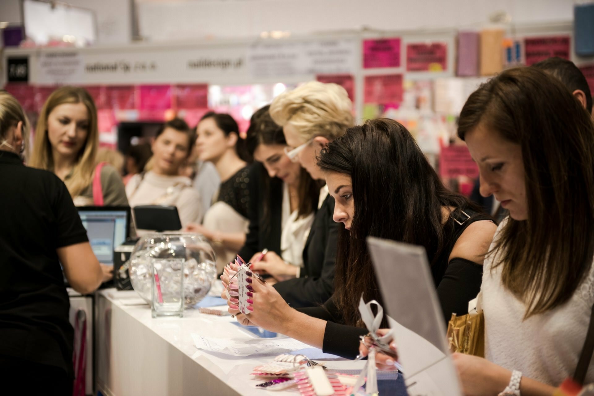 A group of women are standing and sitting at a long table, writing and looking at various things. They look as if they are at an event or workshop captured by event photographer Marcin Krokowski. Various paraphernalia and decorations are on display, some of the women in the background are busy talking, while others are focused on their tasks.  