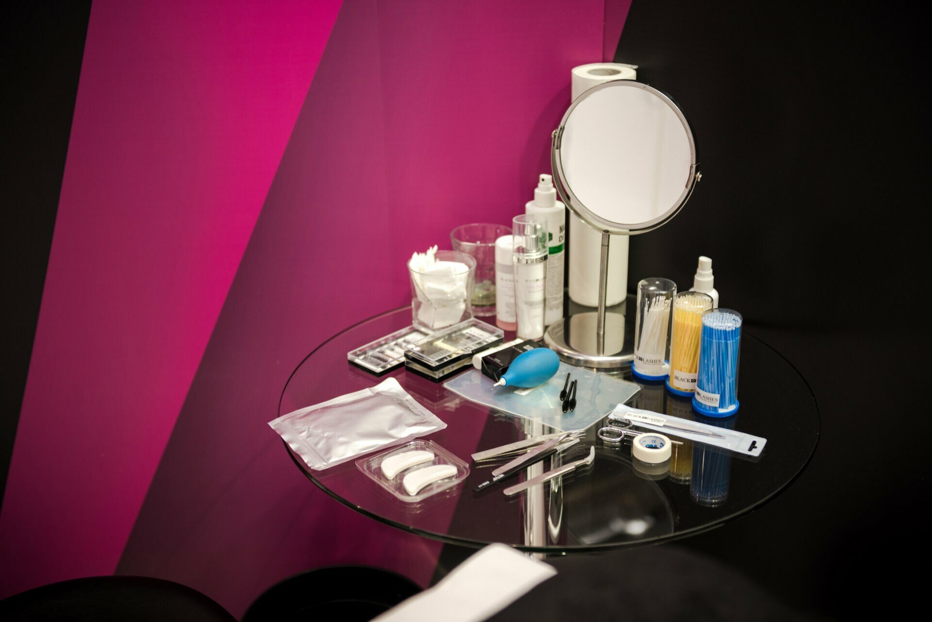 A glass table with various cosmetic and makeup items, including a round mirror, makeup brushes, eyeliners, various containers of cosmetic products, cotton balls and a blue blower. In the background is a pink and black wall - perfectly captured by well-known Warsaw photographer Marcin Krokowski. 