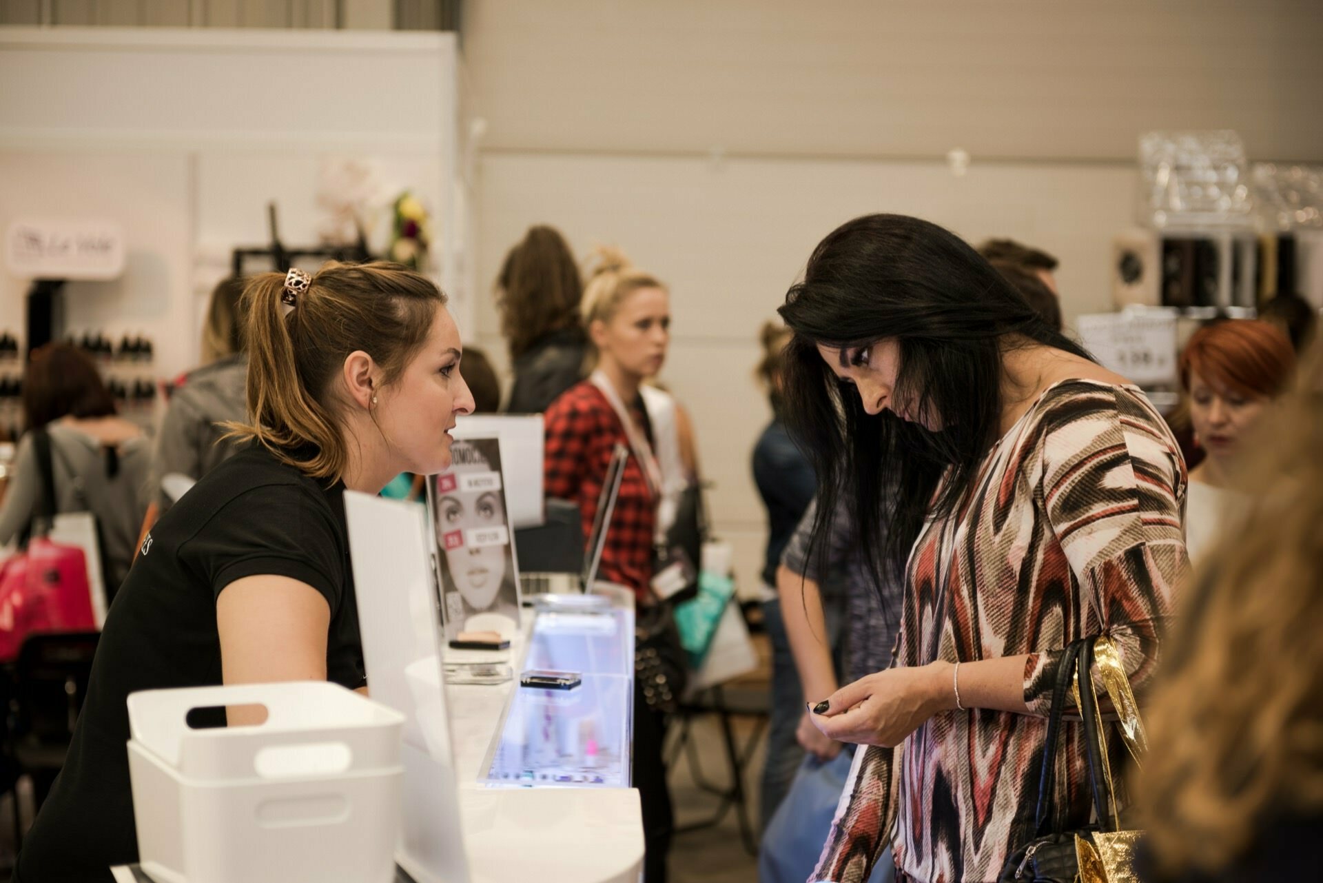 Two women are at a cosmetics stand at a trade fair. One woman behind the counter, probably a representative of Marcin Krokowski - an excellent photographer in Warsaw - is explaining something to the other, who is looking at her phone. In the background you can see other attendees and various booths with cosmetic products.  