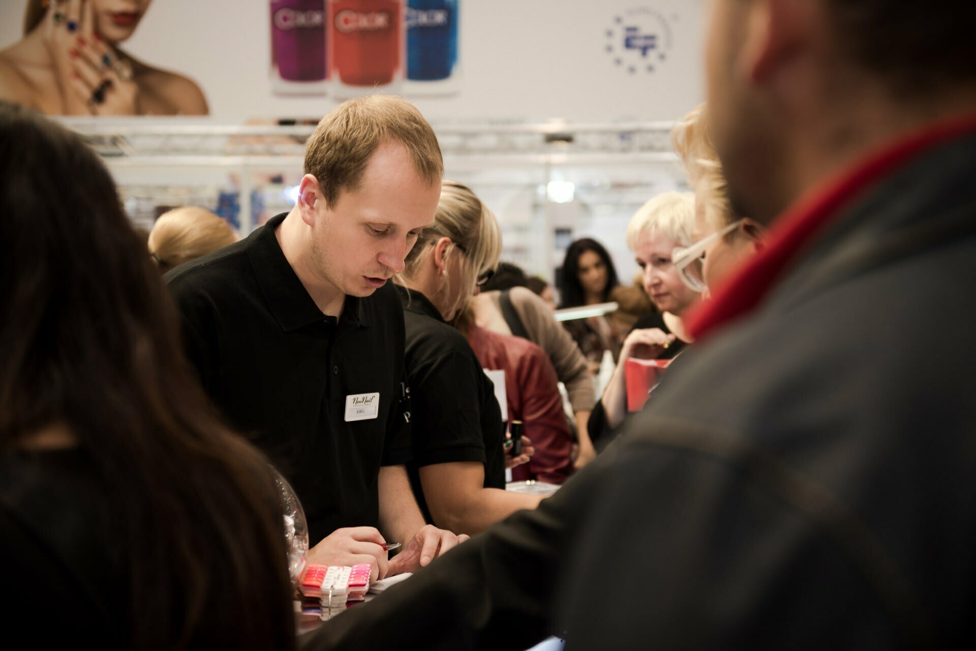 A man with a badge stands behind a counter in a busy exhibition hall and attentively serves customers. Dressed in a black polo shirt, he works diligently while people around him carry bags and carry on conversations. Colorful displays are visible in the background, setting the stage for Marcin Krokowski, a photographer in Warsaw.  