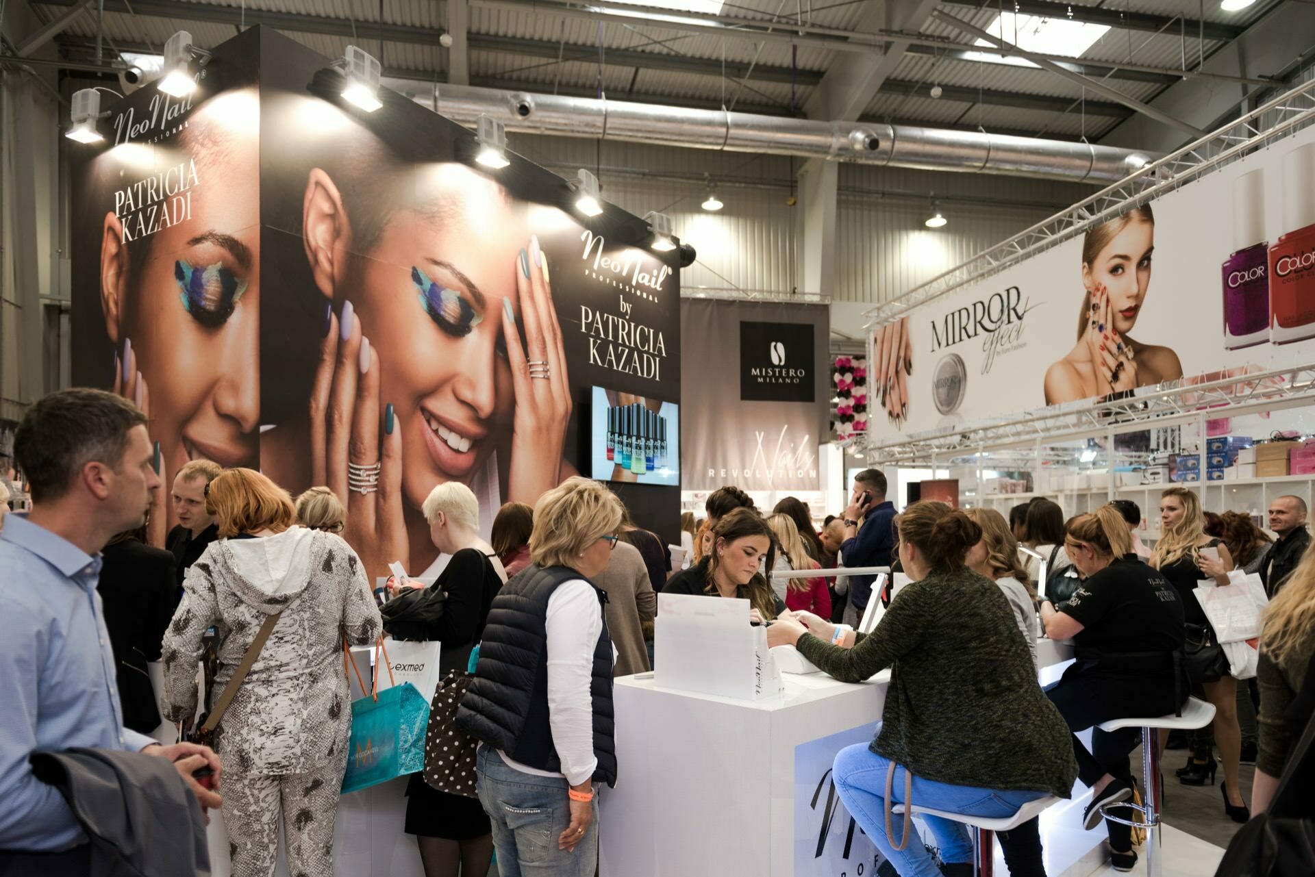 People browsing and interacting at a trade show booth displaying cosmetics and toiletries, elegantly captured by Marcin Krokowski, a well-known photographer in Warsaw. The backdrop is captivating with large banners depicting women in colorful makeup, while product displays and consultation tables are in the foreground. 