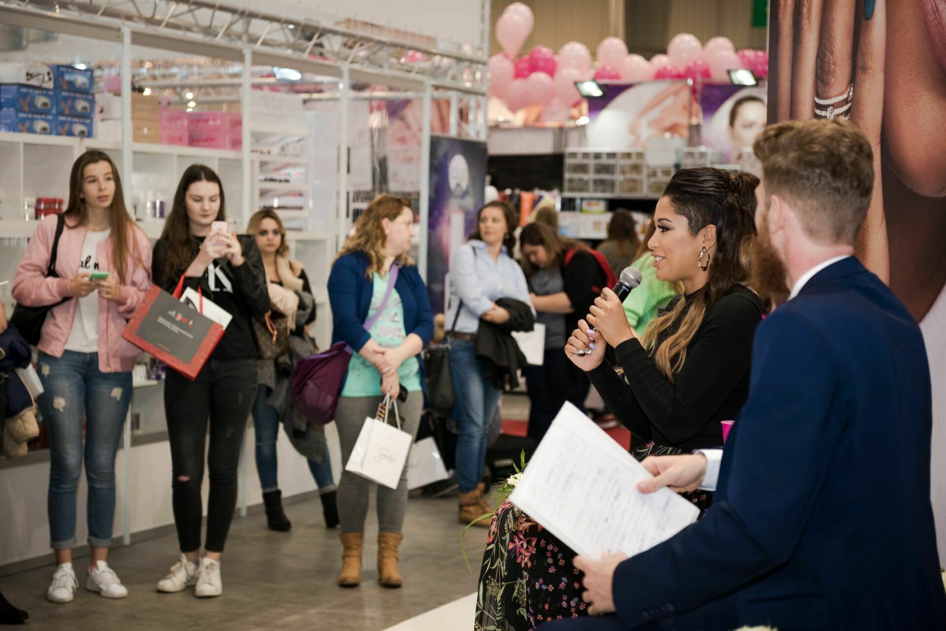 A woman holding a microphone speaks on stage at the event, while a man holding documents sits next to her. A crowd of people stands in front of them and listens. Pink balloons and various product displays are visible in the background, captured by Marcin Krokowski, a well-known event photographer in Warsaw.  