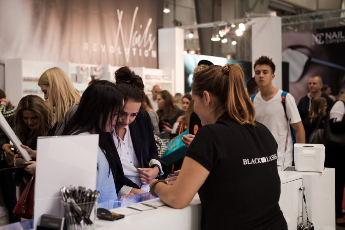 Grupa ludzi gromadzi się wokół stoiska z etykietą „Nails Revolution” podczas ruchliwej konwencji lub targów. Trzy kobiety w skupieniu patrzą na coś na blacie, a pomaga im pracownik, a wszystko to uchwycił Marcin Krokowski, uznany fotograf eventowy. W tle widoczni są inni uczestnicy.