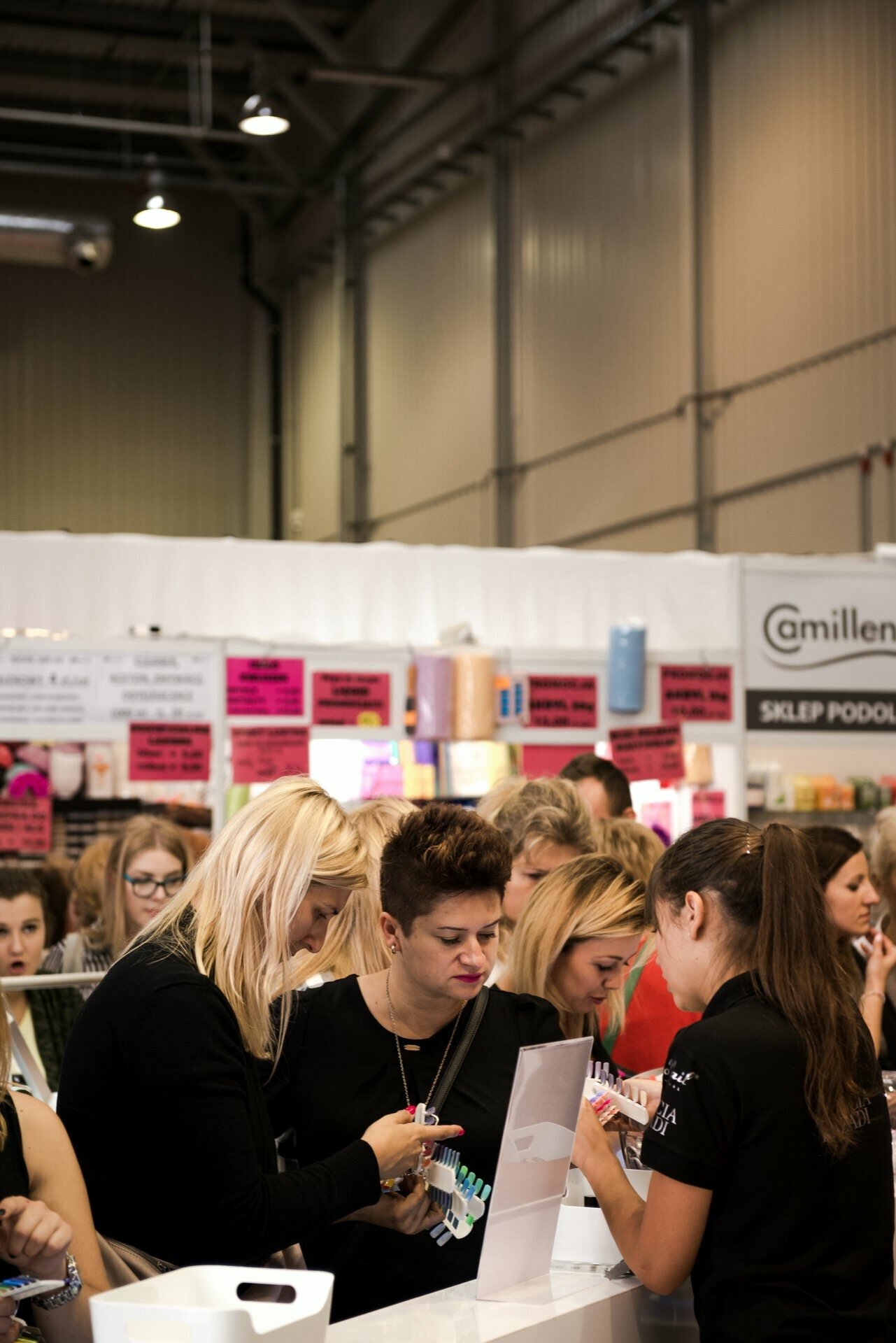 A bustling indoor market captured by Marcin Krokowski, a well-known event photographer in Warsaw. People gather around a booth displaying a variety of products, some examining them closely and others interacting with the booth staff. In the background you can see brightly colored signs and a variety of products.  