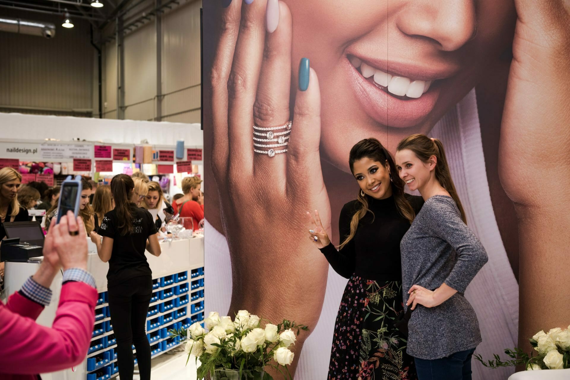 Two women pose for a photo in front of a large advertisement depicting a smiling woman with manicured nails and jewelry. One of the women gives a peace sign. They are in a busy event space, with people and booths in the background, captured by event photographer Marcin Krokowski.  