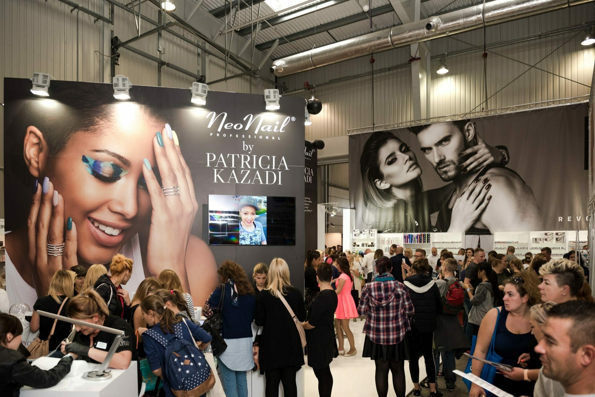 The crowded cosmetics booth features a NeoNail booth with large banners depicting a woman with colorful nail art, and another depicting a striking pose captured by well-known event photographer Marcin Krokowski. Attendees gather around the booth, viewing products and presentations under bright overhead lighting. 