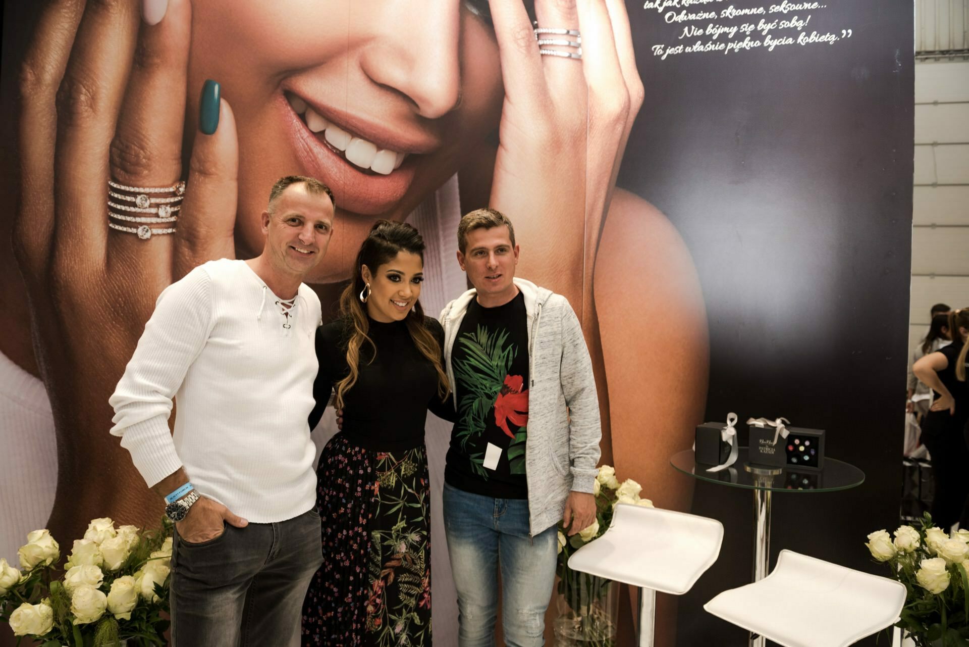 Three people pose in front of a large advertisement showing a close-up of a smiling woman showing rings on her fingers. White roses are arranged in front of the display. To the side are two white chairs and a table with products, captured by Marcin Krokowski, a Warsaw-based event photographer.  