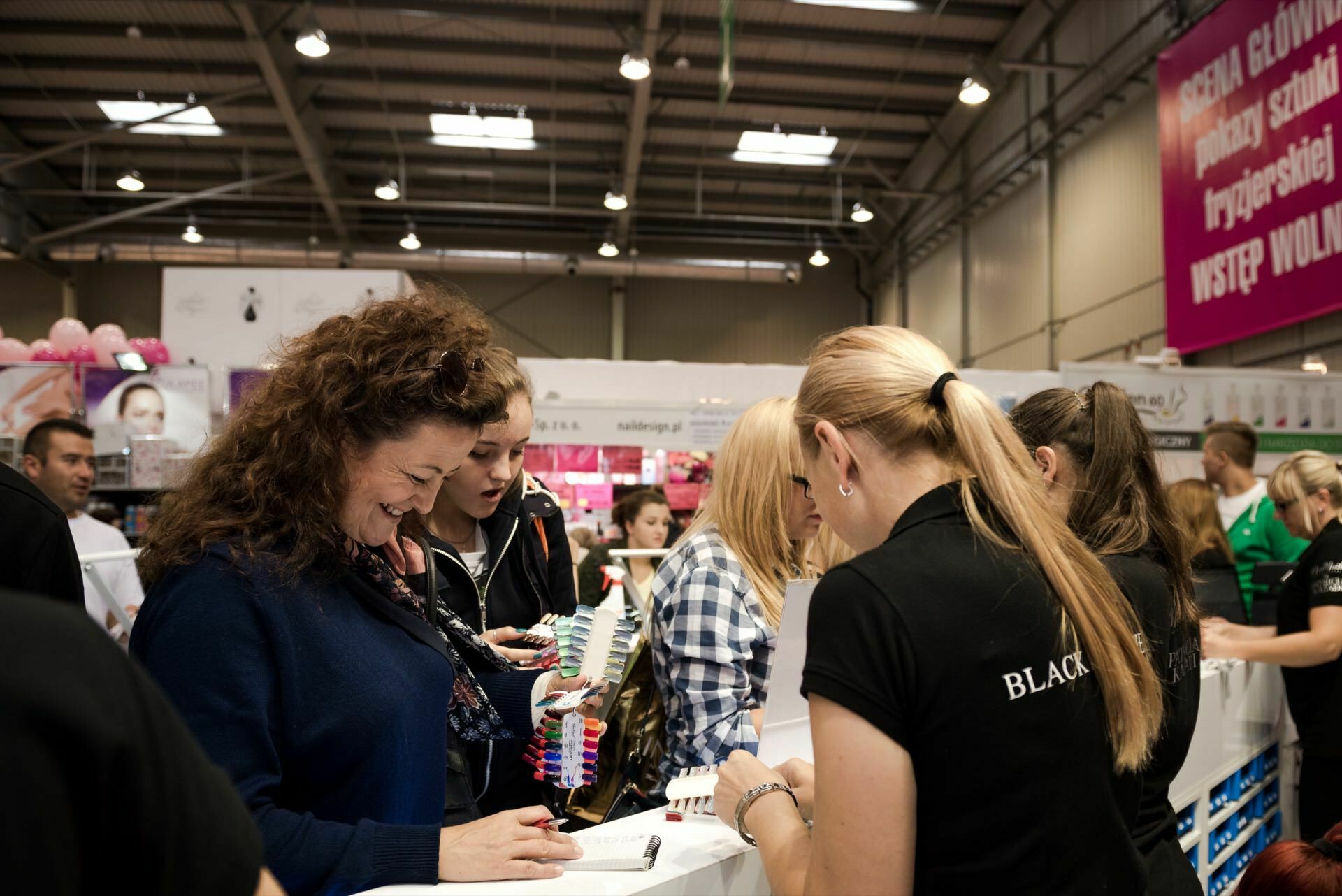 A group of people gathered at a booth during a crowded event held indoors. One woman is smiling and looking at color samples, another is helping her. The premises have high ceilings, with various banners and exhibitions in the background, beautifully captured by Marcin Krokowski, a well-known photographer in Warsaw.  