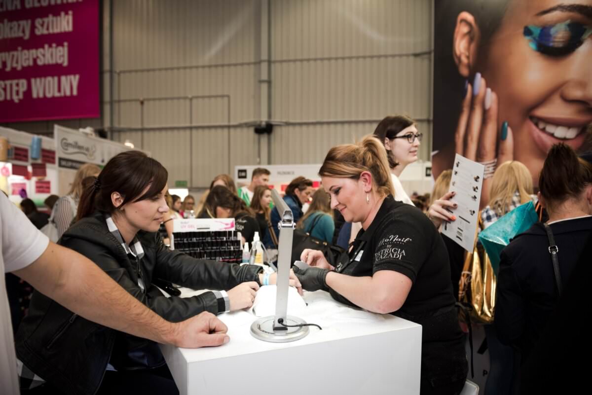Kobieta robi manicure przy stoisku podczas pracowitego wydarzenia. Manikiurzystka pilnie pracuje nad paznokciami, podczas gdy w tle krzątają się ludzie. Pobliska reklama zdobi dużą uśmiechniętą twarz, a nad nią widać duży różowy szyld z tekstem w języku polskim – doskonale uchwycił fotograf eventowy Marcin Krokowski.