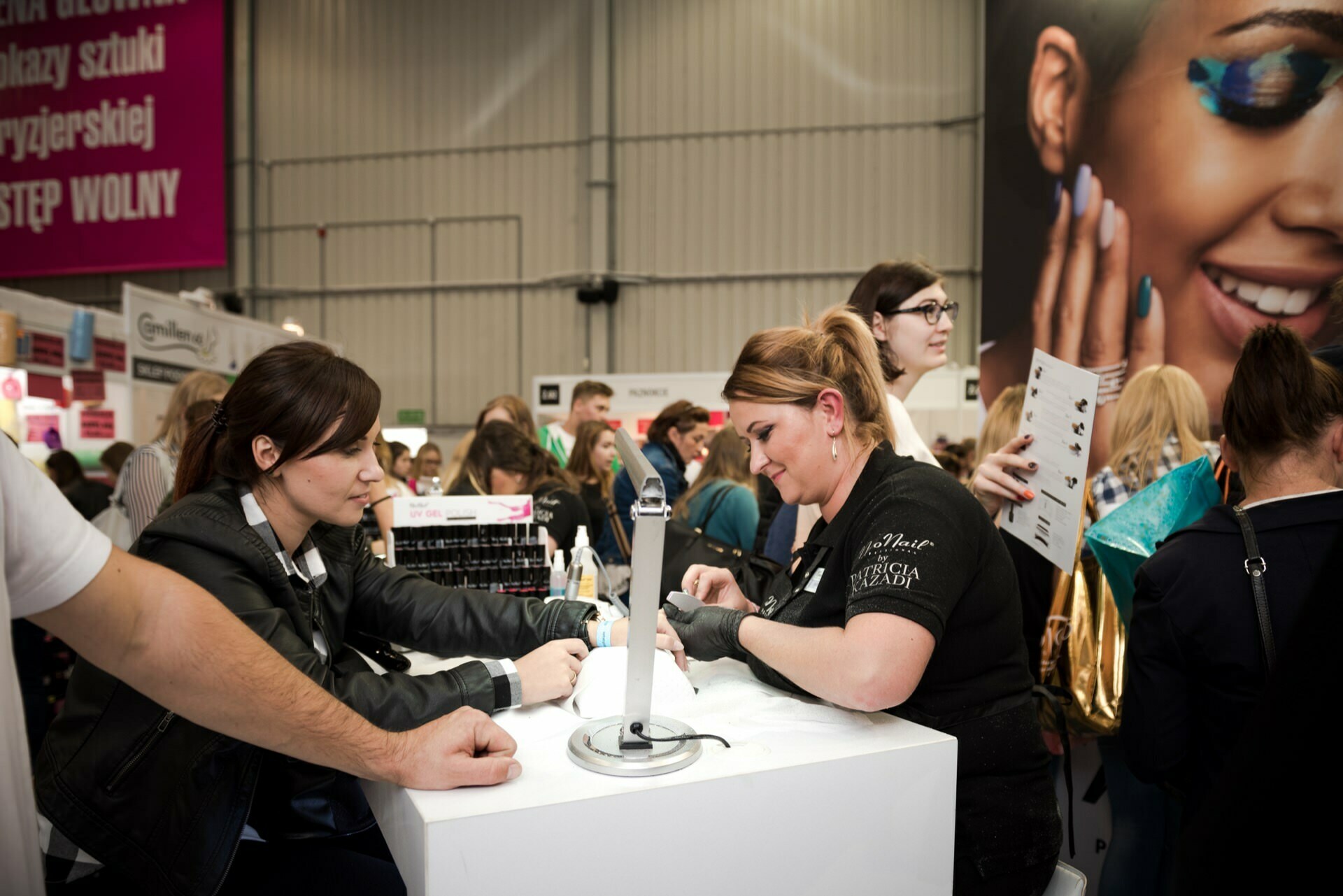 A woman gets a manicure at a booth during a busy event. The manicurist works diligently on her nails while people shuffle around in the background. A nearby advertisement adorns a large smiling face, and a large pink signboard with text in Polish can be seen above it - perfectly captured by event photographer Marcin Krokowski.  