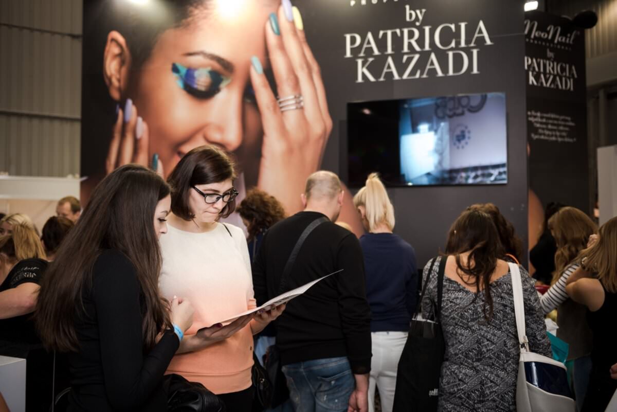 Tłum gromadzi się na stoisku wystawowym z widocznym napisem „autor: PATRICIA KAZADI”. Dwie kobiety na pierwszym planie uważnie przyglądają się broszurze, a stoisko zdobi duży ekran i wizerunek kobiety z makijażem i biżuterią, uchwycony przez Marcina Krokowskiego, fotografa w Warszawie.