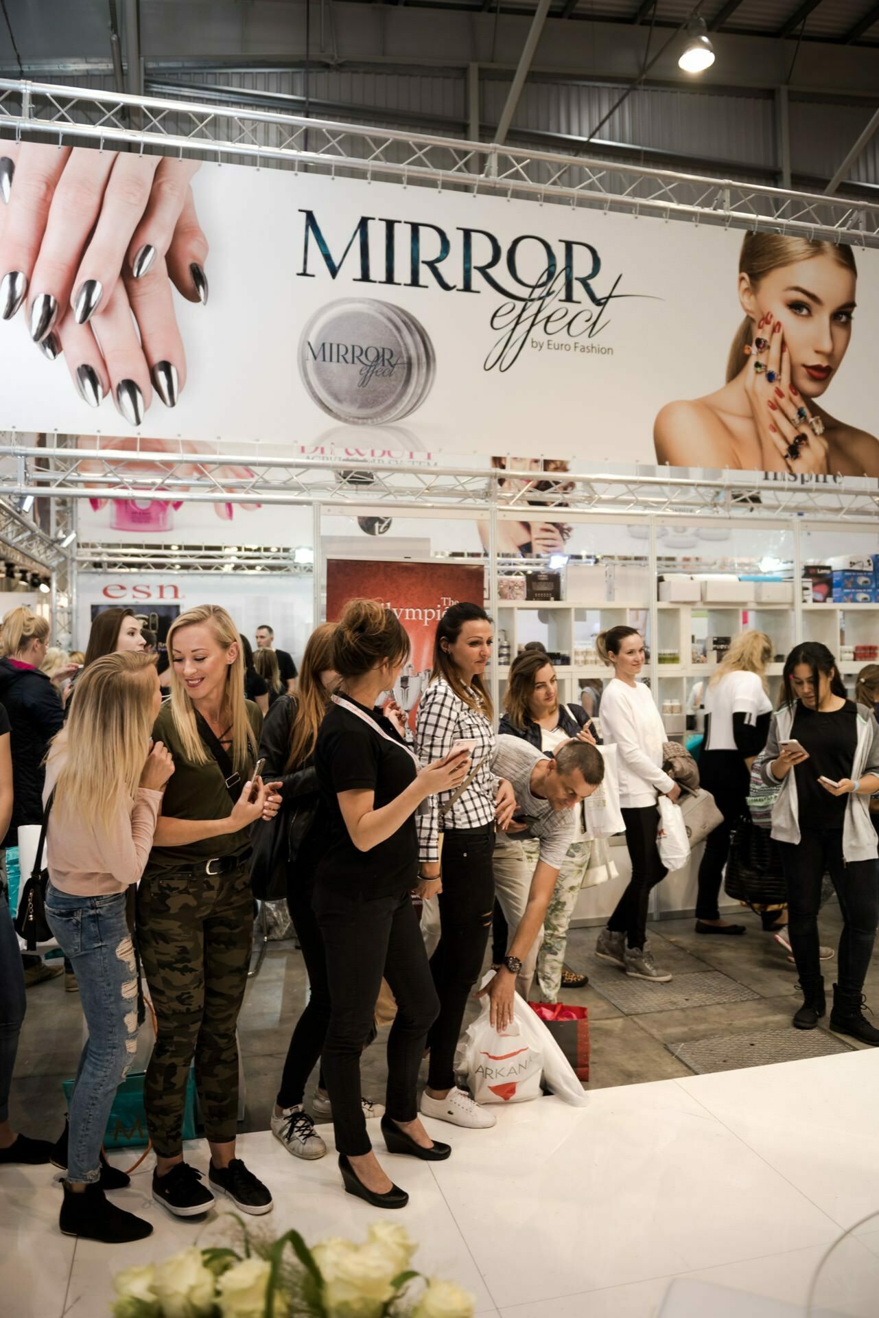 A group of people are standing in line at a cosmetics fair. Above them, a large advertisement showing perfectly manicured nails and a model with makeup, promoting the "Mirror Effect" brand Euro Fashion. In the background, booths with various cosmetic products are visible, captured by event photographer Marcin Krokowski.  