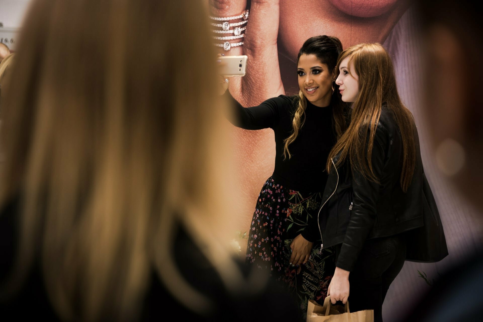 Two women take a selfie together in a room. One has dark hair, is wearing a black top and floral skirt; the other has light brown hair, a black jacket and is holding a shopping bag. They are standing in front of a large display, which features a close-up of their faces taken by Marcin Krokowski, a talented photographer in Warsaw.  