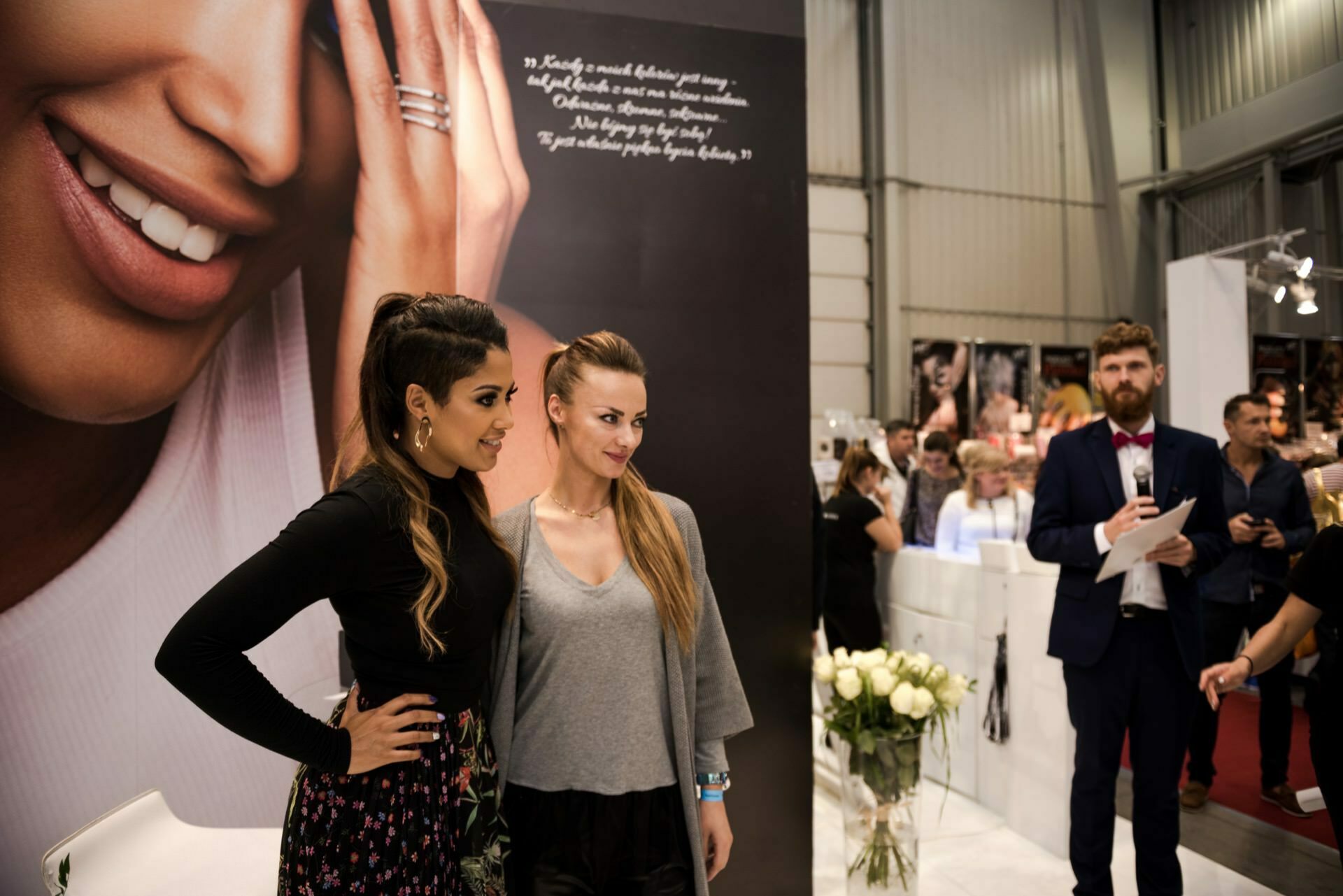 Two women pose for a photo in front of a large poster depicting a woman's face. The background appears to be part of a trade fair or exhibition. A crowd can be seen in the background, and Marcin Krokowski, a well-known photographer in Warsaw, is holding the microphone.  