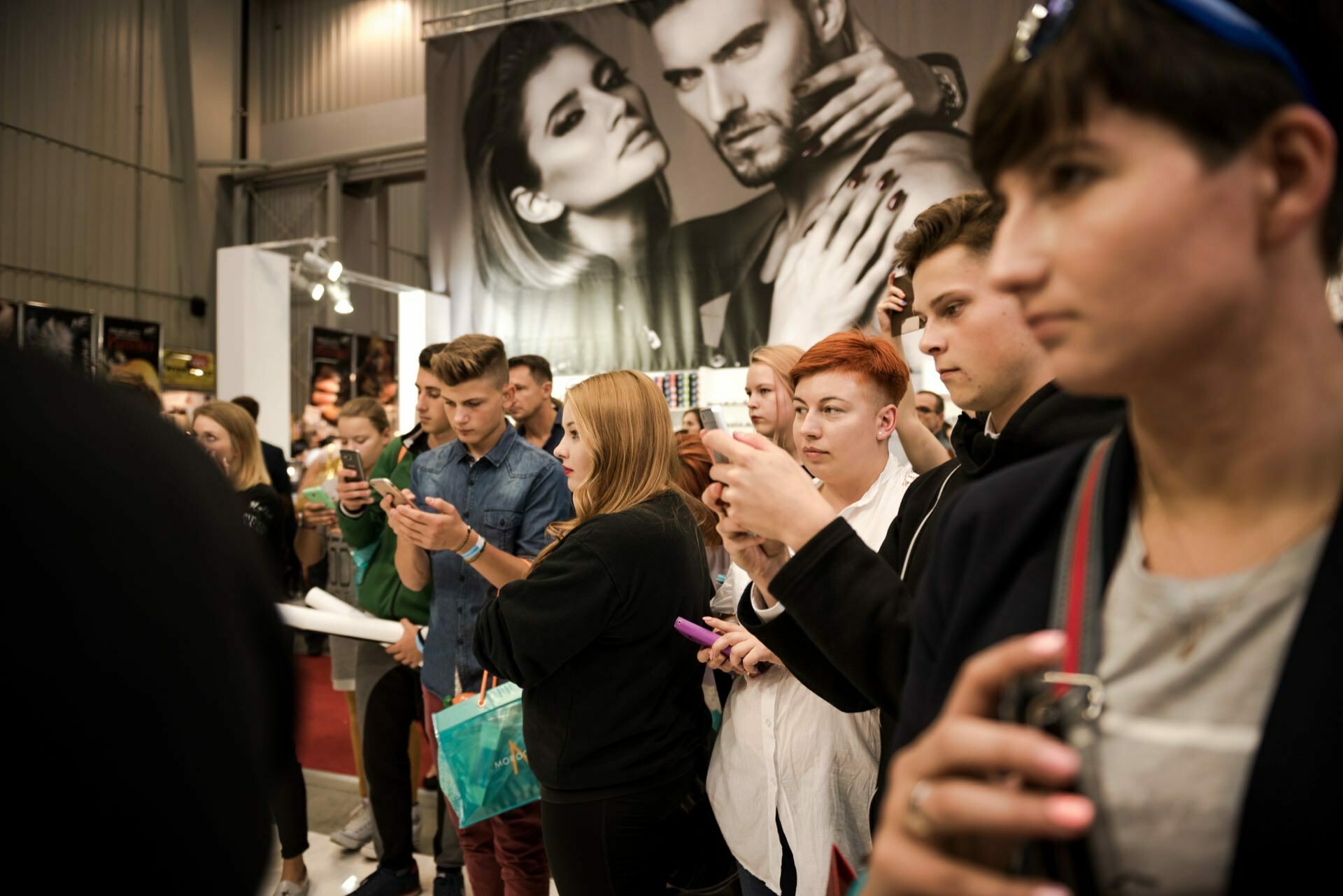 A group of people standing close together, many of them holding their phones up, presumably taking photos or videos. In the background is a large poster with an image of a man and woman posing. The scenery appears to be a closed event, captured by Marcin Krokowski, a well-known photographer in Warsaw.  