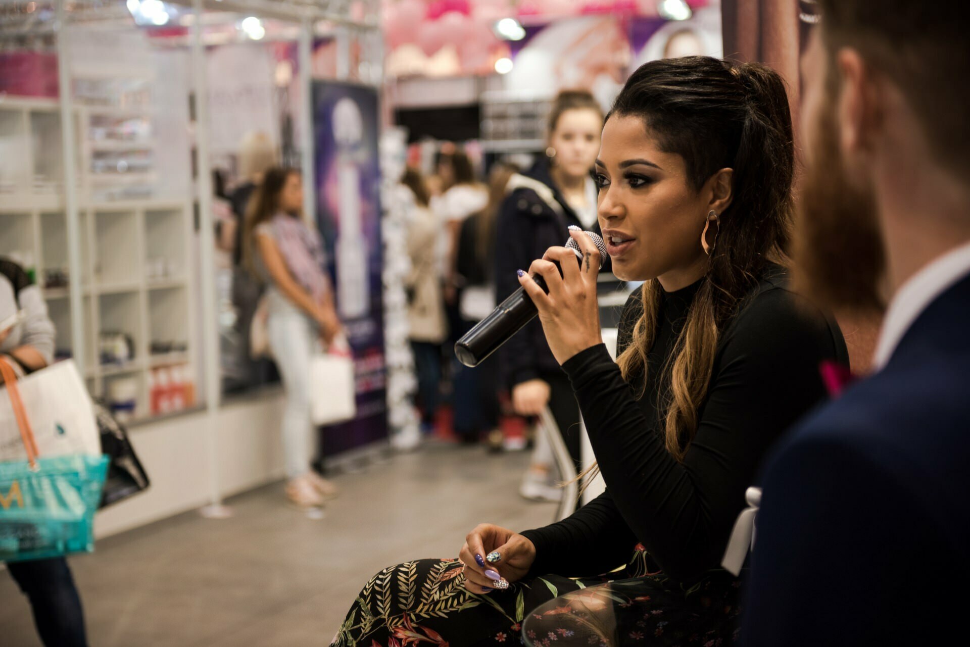 A woman with long, dark hair speaks into a microphone while seated. She is dressed in black with floral patterns. In a blurry background captured by well-known Warsaw photographer Marcin Krokowski, people walk and store in a store with shelves of products and promotional displays.  