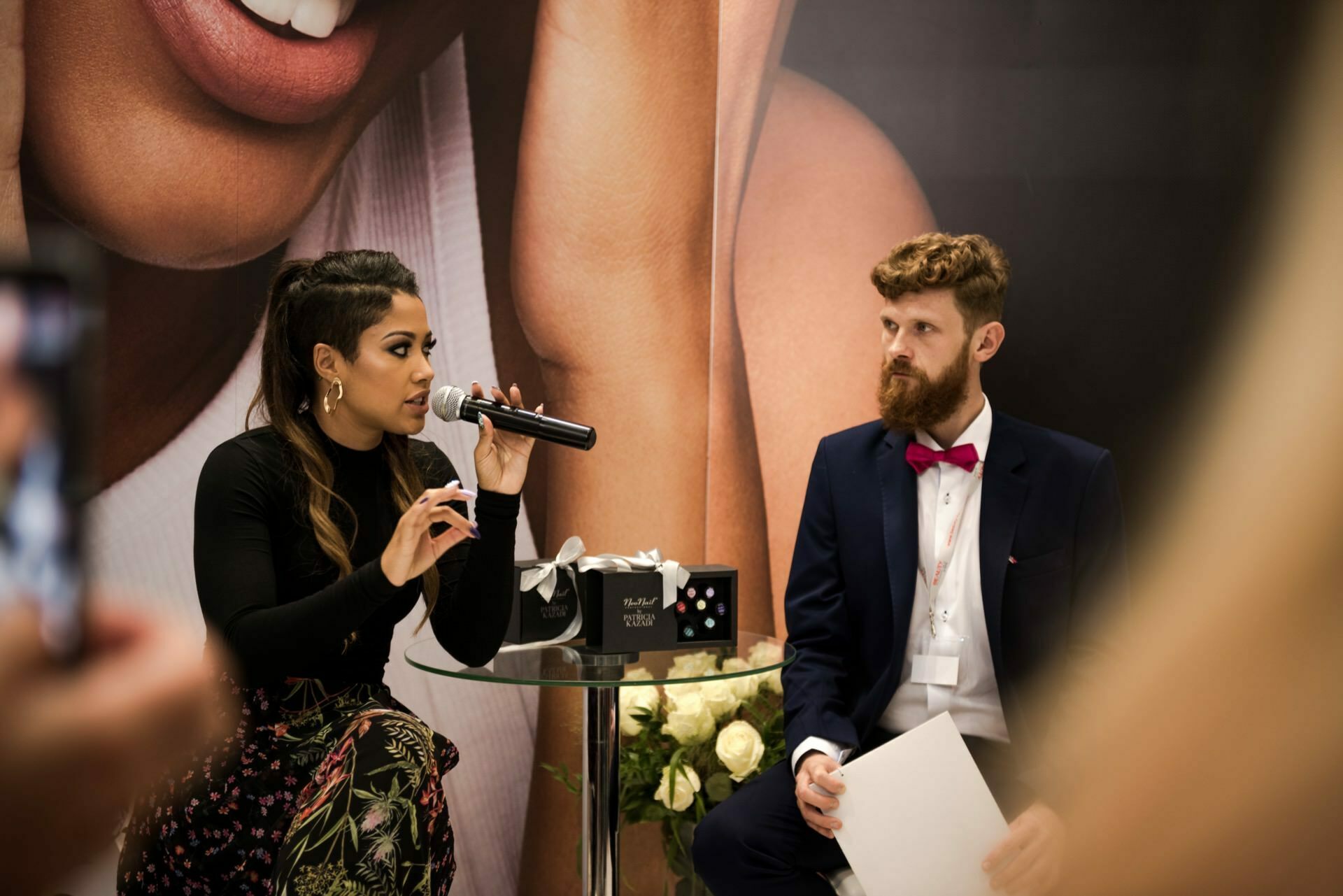 The woman on the left, holding a microphone, speaks passionately, while the man with a beard and bow tie on the right listens attentively, holding a piece of paper. They sit at a small glass table, against a backdrop of a bouquet of white roses. The moment is captured by Marcin Krokowski, a photographer in Warsaw.  