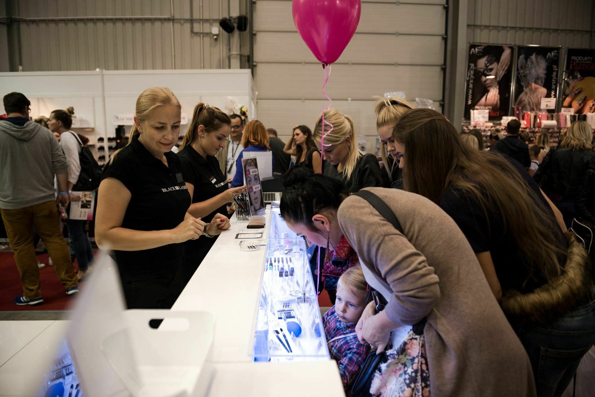 People gather around a booth at the fair. A blonde woman in a black shirt stands behind the counter and talks to the attendees. A child looks out from over the counter, carefully examining the exhibition. In the background, photographer Marcin Krokowski, known in Warsaw as a prominent event photographer, captures the moment when someone holds a pink balloon.   