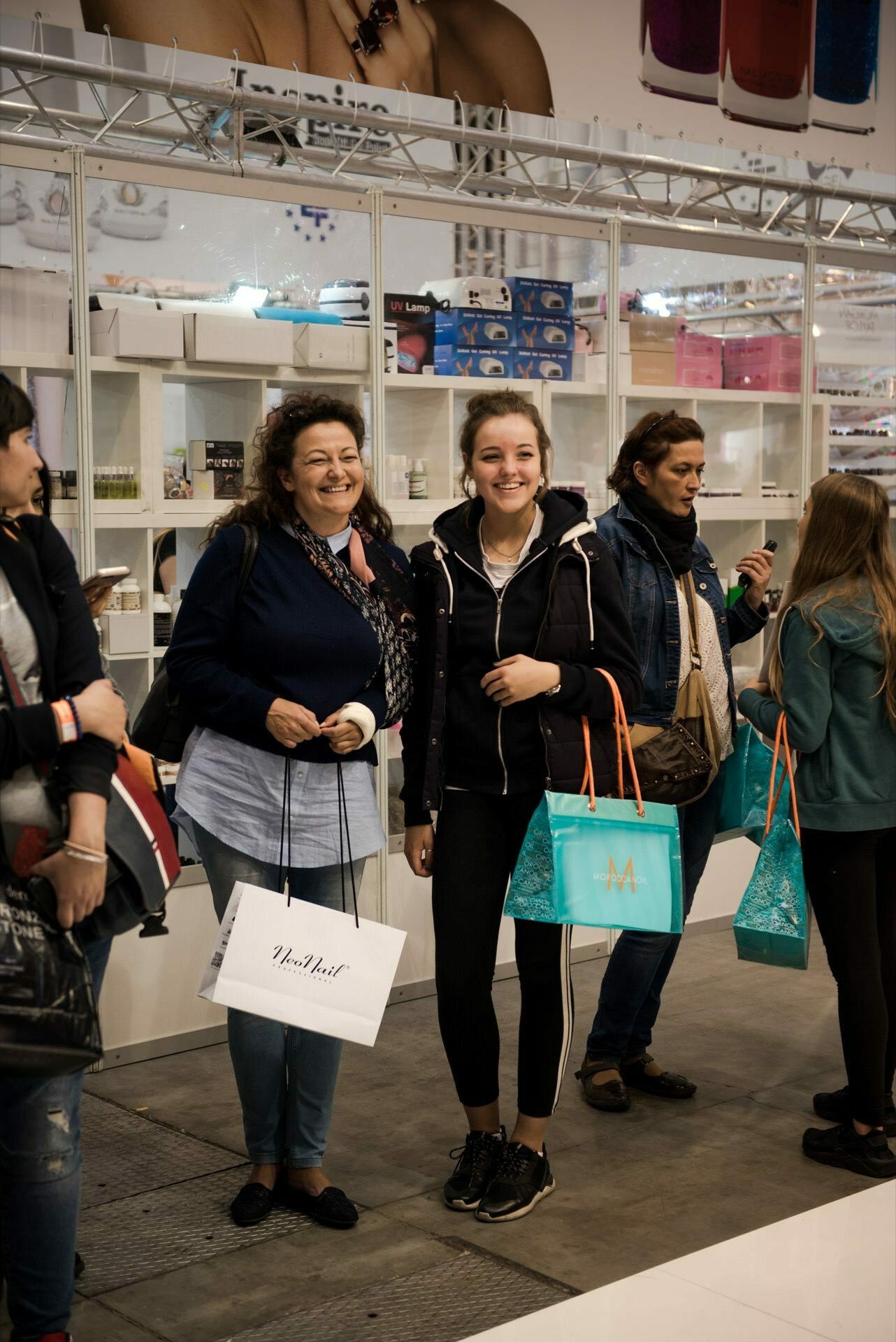 A group of people stand in front of a stand at a fair or market. Two women in the foreground, captured by Marcin Krokowski, are smiling and holding shopping bags. The background shows various products on display, and other people can be seen browsing the booth in the background.  