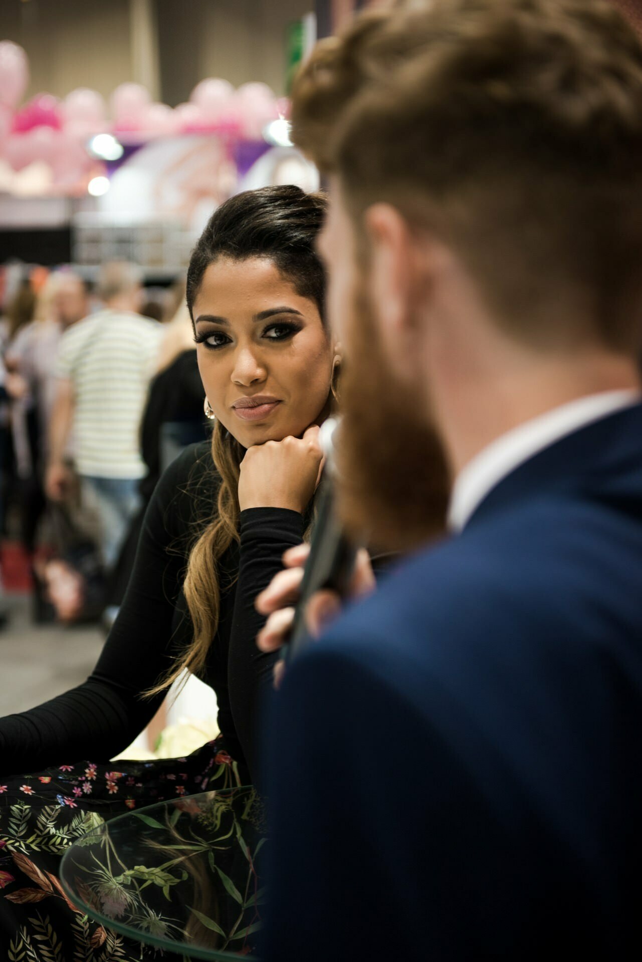 A woman with long hair and hoop earrings listens intently to a bearded man speaking into a microphone. They are in a crowded room that looks like an event or conference. She is wearing a floral skirt and black top, which was perfectly captured by photographer in Warsaw, Marcin Krokowski.  