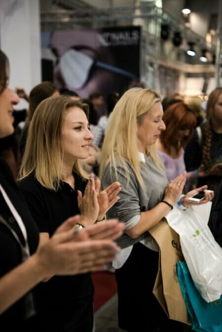 Grupa klaszczących i uśmiechających się ludzi na imprezie w hali, doskonale uchwycona przez fotografa eventowego Marcina Krokowskiego. Są ubrani w zwykłe ubrania i trzymają przedmioty takie jak torby i telefony. W tle stoisko z widoczną częścią szyldu z napisem „NAILS.