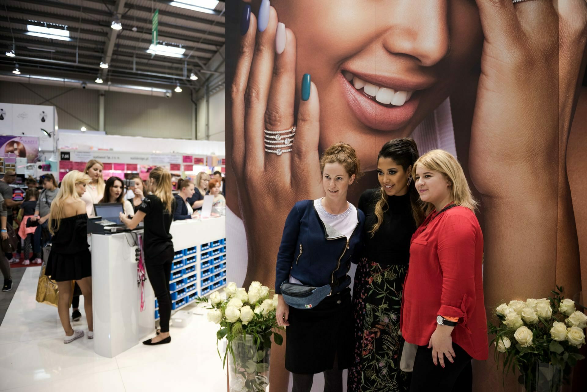 Three women pose together for a photo by prominent Warsaw photographer Marcin Krokowski in front of a large advertisement depicting a smiling woman with manicured nails. Behind them is a bustling exhibition space with a mix of people and booths displaying a variety of products. 