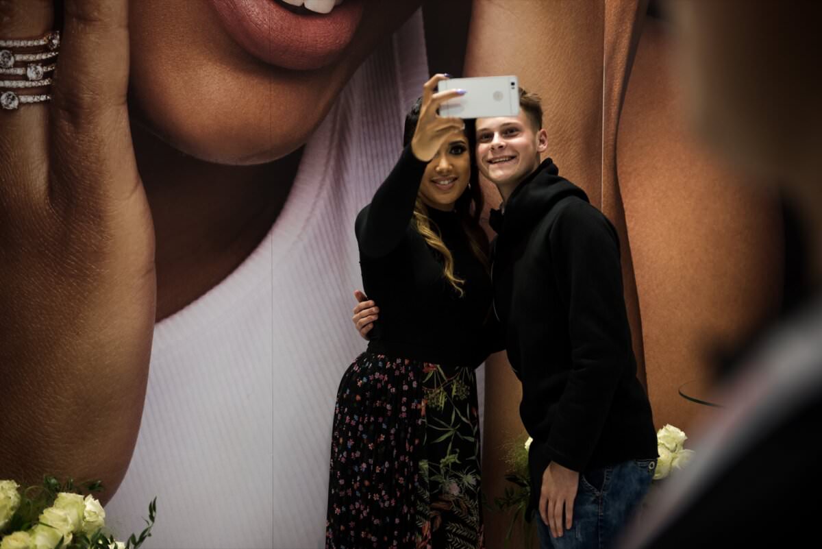 Dwie osoby pozują do selfie przed dużym plakatem reklamowym przedstawiającym zbliżenie twarzy. Osoba po lewej stronie, prawdopodobnie inspirowana standardami fotografa w Warszawie, trzyma telefon i robi zdjęcie. Obie osoby uśmiechają się i wydają się cieszyć tą chwilą. W pobliżu znajdują się białe kwiaty.