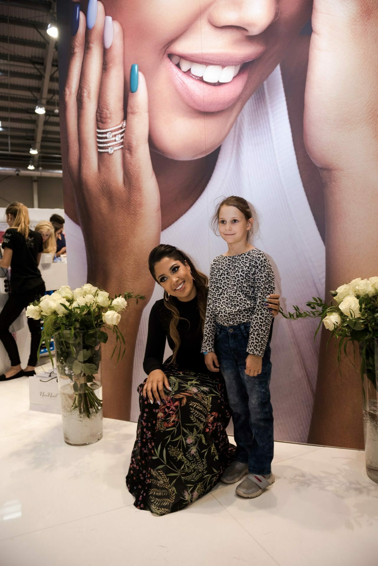 A woman crouches and smiles next to a young girl standing, both of them posing in front of a large poster depicting a smiling face and a manicured hand. On either side of them stand vases of white roses. The background appears to be a closed event or exhibition captured by a well-known photographer in Warsaw, Marcin Krokowski.  