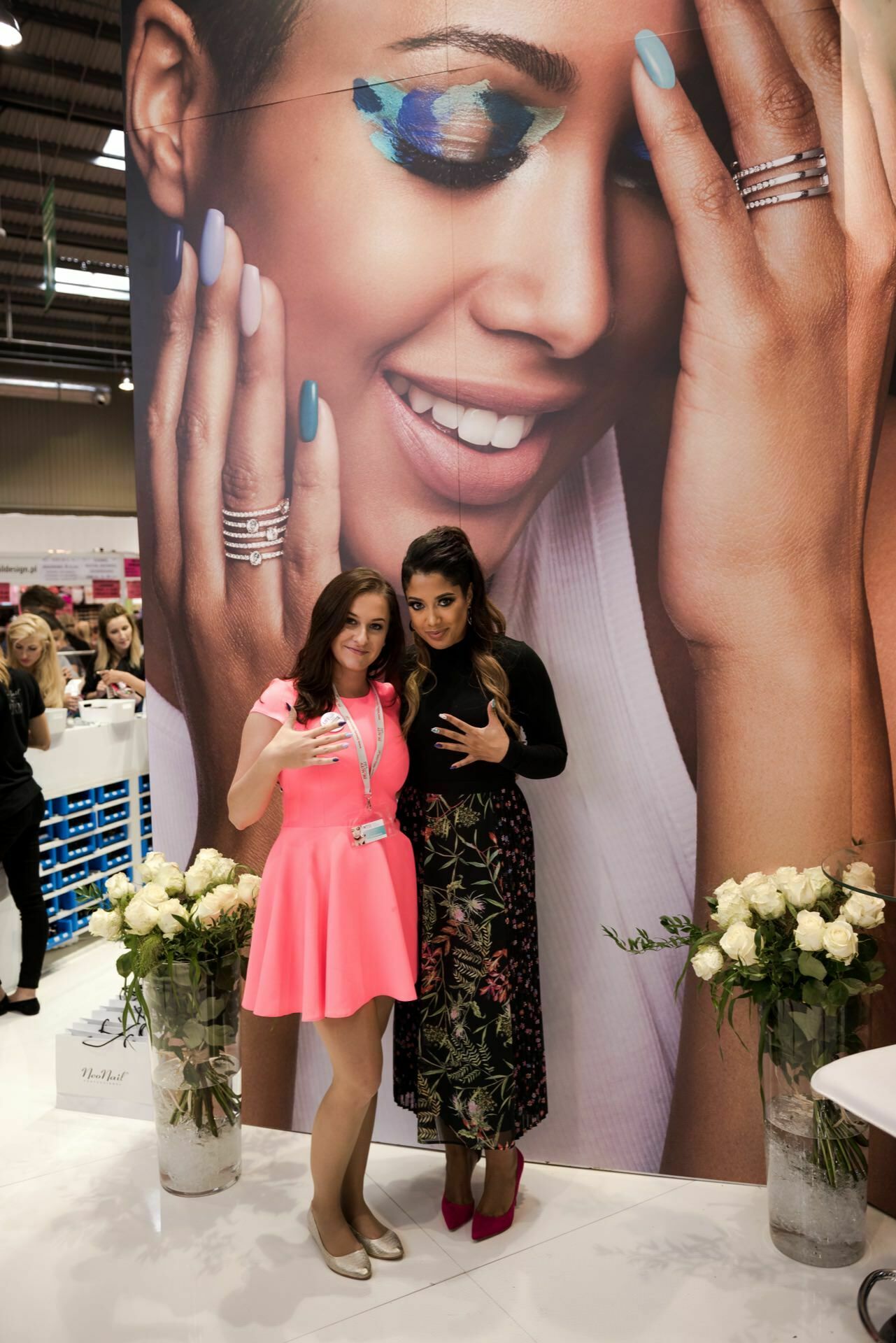 Two women pose together in front of a large promotional poster showing a woman with colorful eye shadow and manicured nails presenting jewelry. This moment was captured by Marcin Krokowski, an acclaimed photographer in Warsaw. The woman on the left is wearing a bright pink dress and a lanyard, while the woman on the right is wearing a black top and floral skirt. On either side stand two vases with white roses   