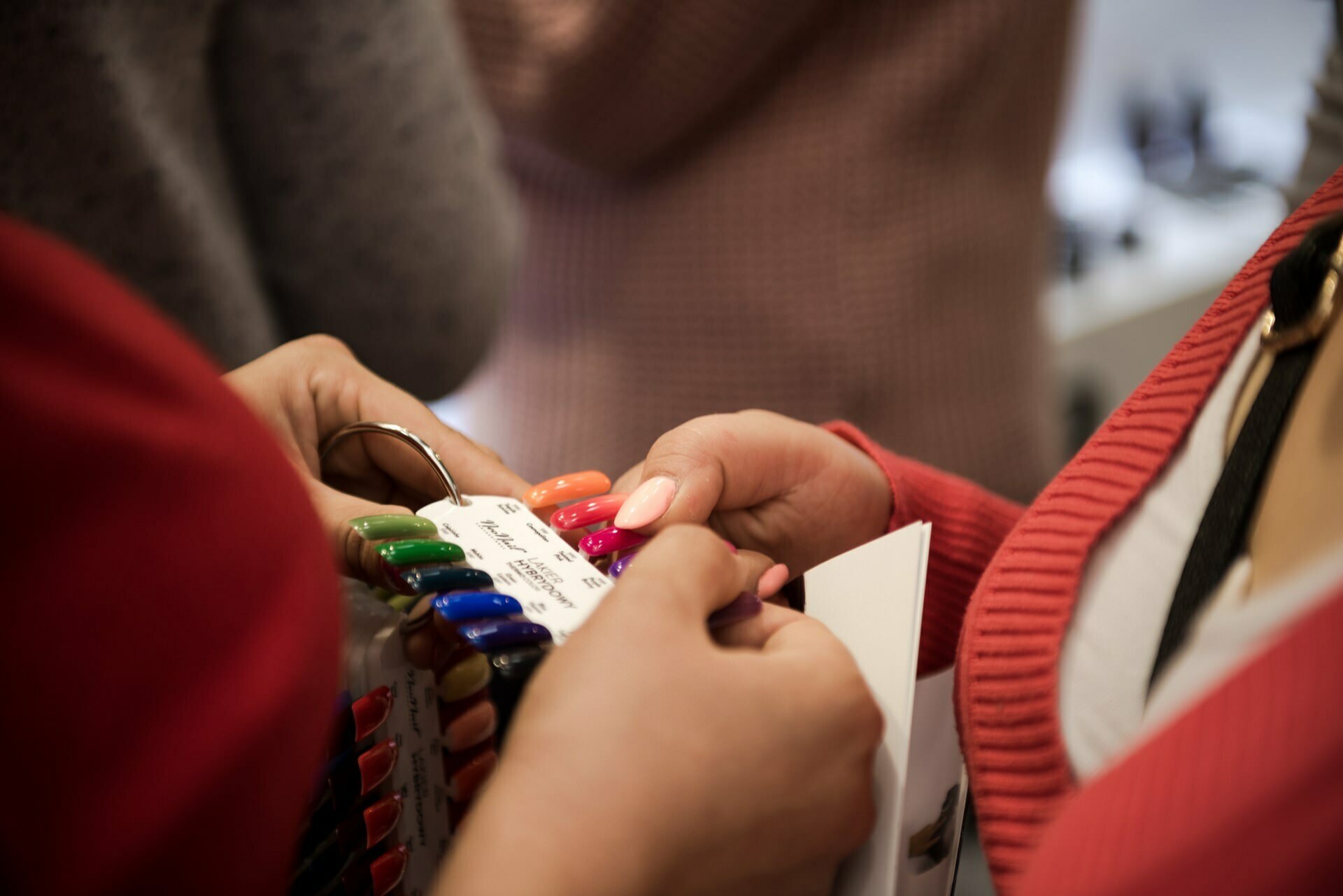 Two people choose a nail polish color from a sample ring in a photograph by Marcin Krokowski. One person holds the ring, showing an array of vibrant color swatches, from green and blue to pink and orange. The background is blurred, focusing attention on the hands and the color samples.  