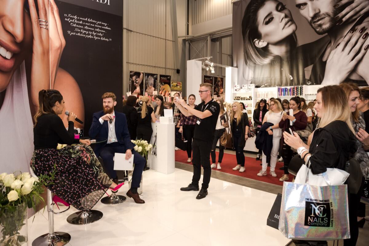 Mężczyzna i kobieta siedzą na stołkach z mikrofonami i rozmawiają z publicznością na konwencji Beauty Forum. Za nimi znajdują się duże plakaty z modelkami. Uczestnicy stoją wokół, niektórzy robią zdjęcia telefonami, inni trzymają torby z upominkami. Otoczenie jest nowoczesne i dobrze oświetlone.