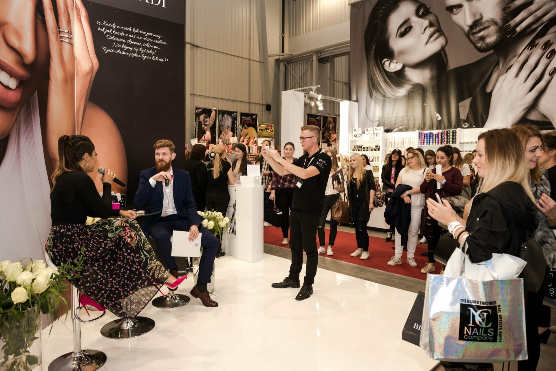 A man and a woman sit on stools with microphones and talk to the audience at the Beauty Forum convention. Behind them are large posters with models. Attendees stand around, some taking pictures with their phones, others holding gift bags. The environment is modern and well-lit.   