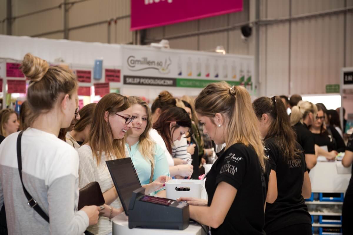 Grupa ludzi zebrała się na stoisku targowym wewnątrz dużego budynku w stylu industrialnym. Niektóre osoby nawiązują kontakt z wystawcami za ladą, inne czekają w tle. Stoisko, udokumentowane przez Marcina Krokowskiego, fotografa eventowego w Warszawie, wyposażone jest w ekrany komputerowe i materiały promocyjne.