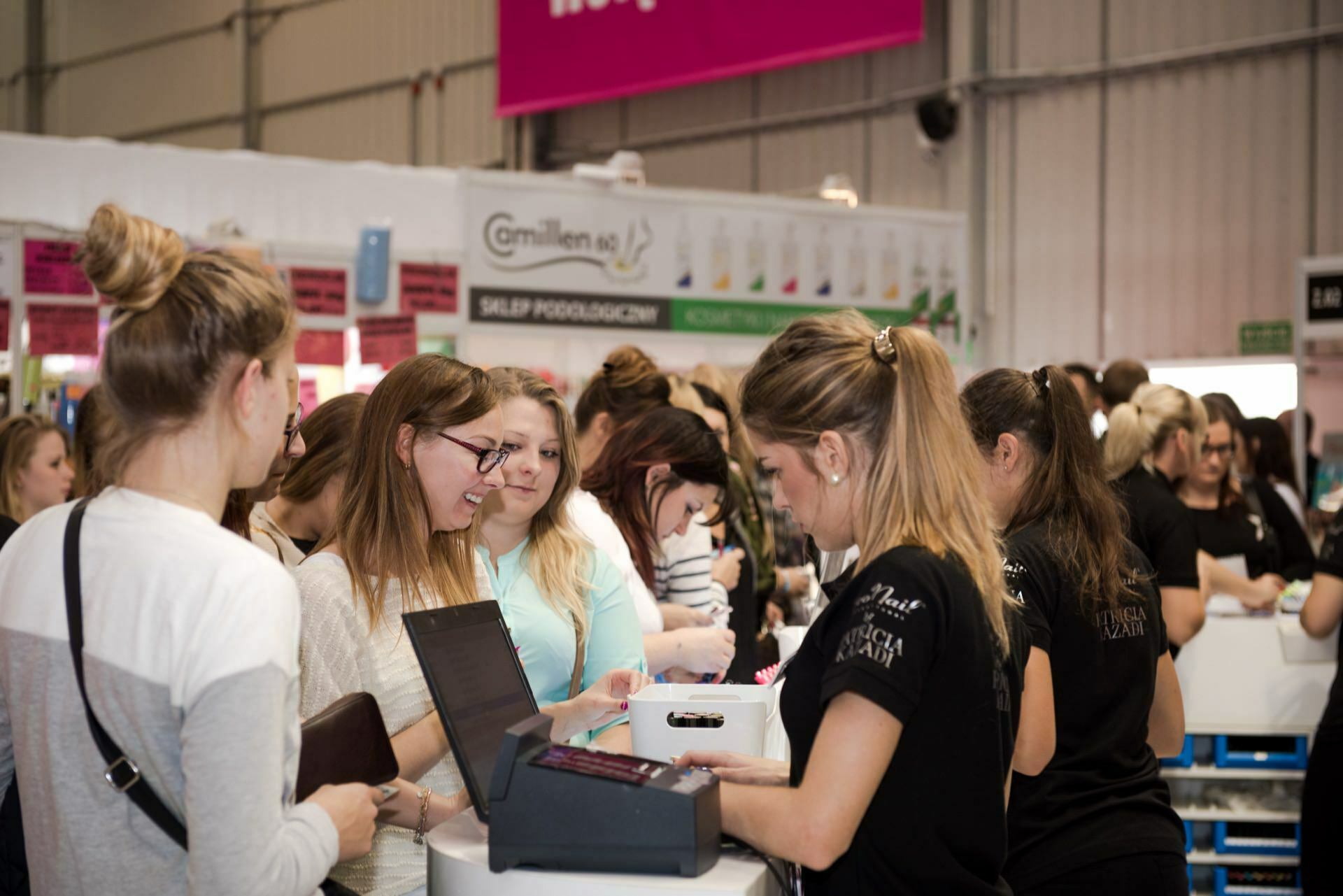 A group of people have gathered at a trade show booth inside a large industrial-style building. Some people interact with exhibitors behind the counter, while others wait in the background. The booth, documented by Marcin Krokowski, an event photographer in Warsaw, is equipped with computer screens and promotional materials.  