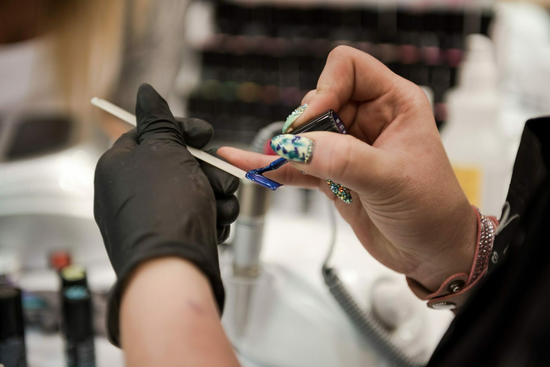 The close-up shot by Marcin Krokowski shows a person applying dark blue nail polish to a wooden stick with a brush, demonstrating nail decoration tools and techniques. The person is wearing a black glove on one side and colorful nail decorations on the other, with smudged nail polish bottles in the background. 