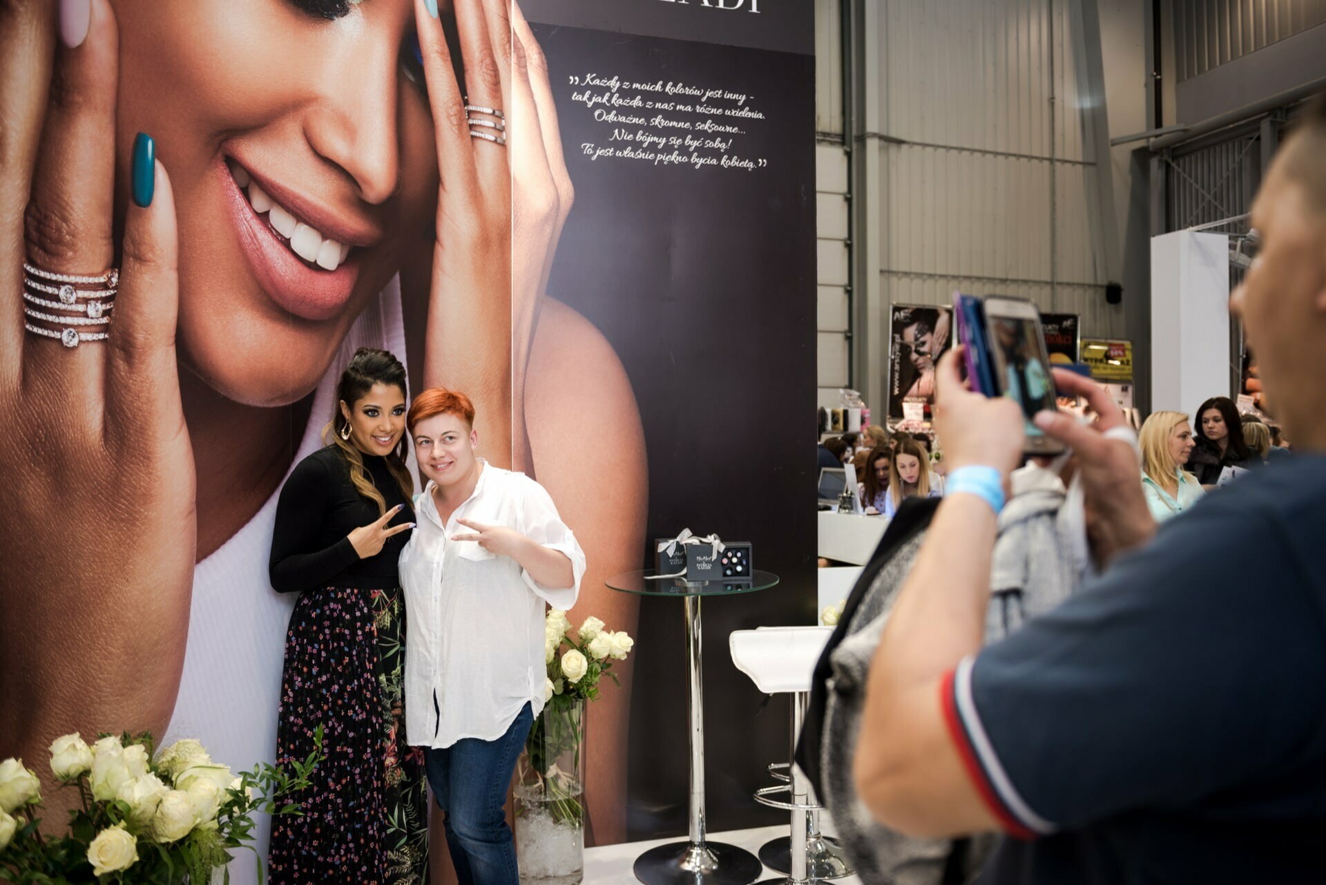 Two women stand together in front of an exhibitor's booth with a large advertisement showing a model displaying jewelry. One person, holding a camera phone, takes a picture of them, capturing the moment for Marcin Krokowski, a well-known event photographer in Warsaw. The booth is decorated with white roses, amid a busy background full of people.  