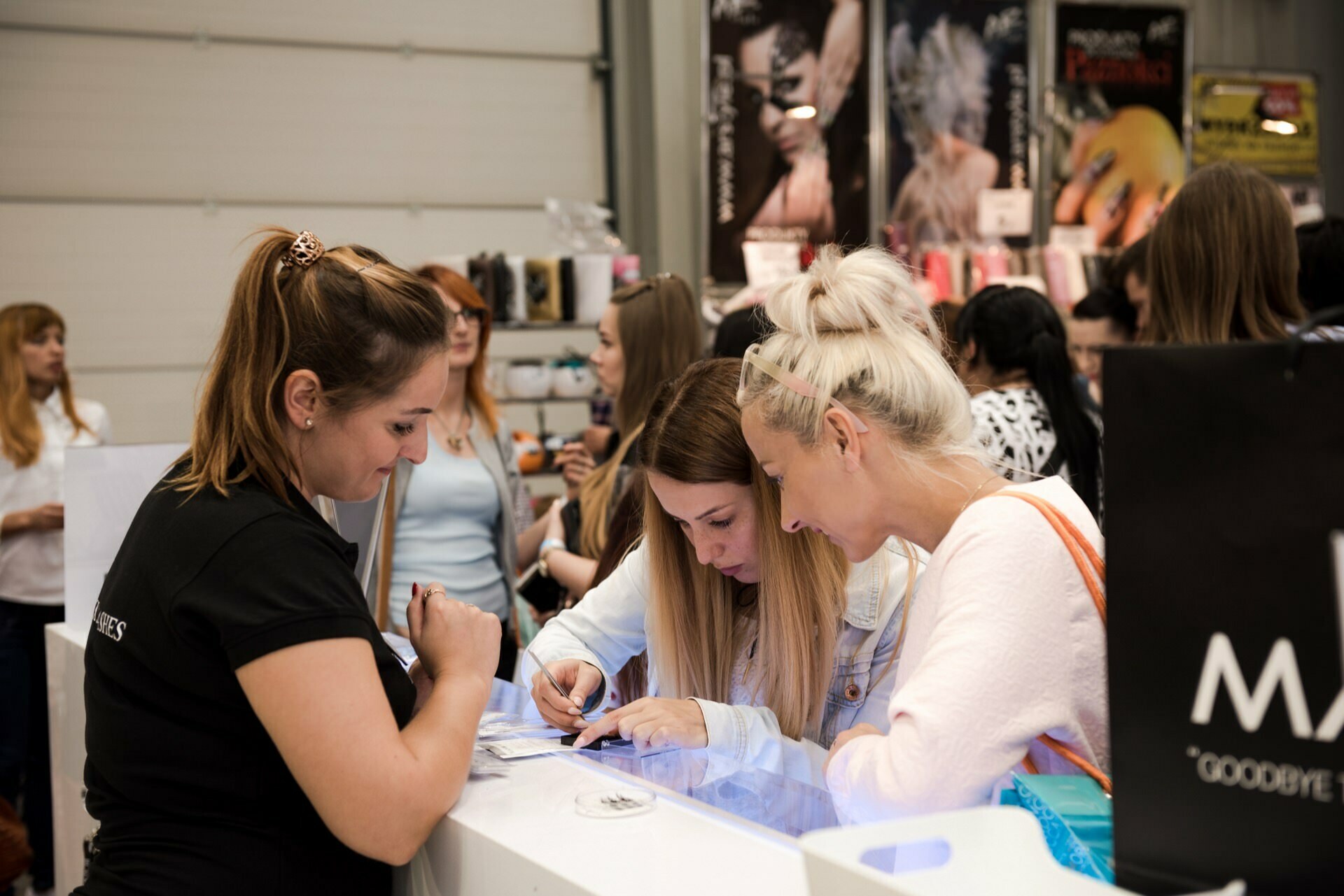 A group of people gather around a booth at a convention or trade show. Three women in the foreground are focused on filling out forms on the counter, while others in the background are browsing various products and displays. The atmosphere is vibrant, and Marcin Krokowski captures every moment as a photographer of the event.  