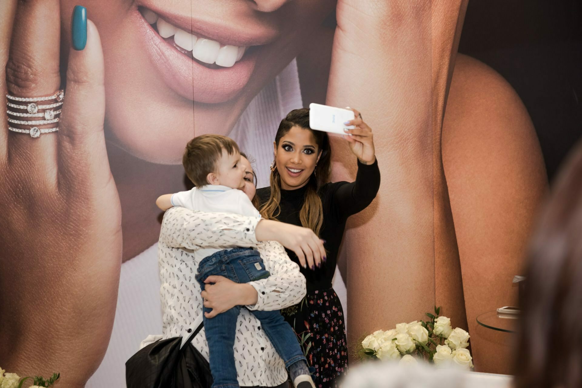 A woman holding a small child while taking a selfie, both smiling at a smartphone screen. Behind them is a large advertisement showing the woman's smiling face and jewelry. Next to them on a table is a vase of white roses, perfectly captured by Marcin Krokowski, a photographer in Warsaw.  