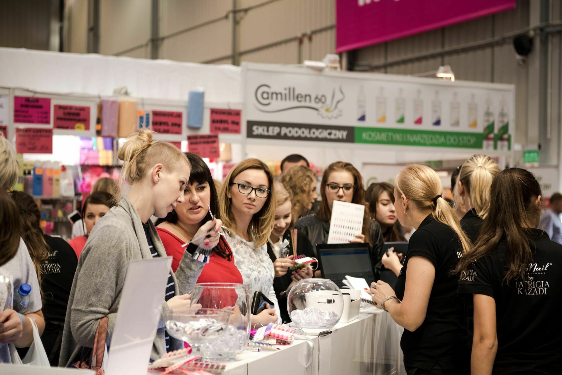 A bustling scene at the fair with several people gathered around the booth. The booth is decorated with various signs and products, and the staff is busy chatting and handling transactions. Attendees, captured by event photographer Marcin Krokowski, seem interested and look at the items on display.  