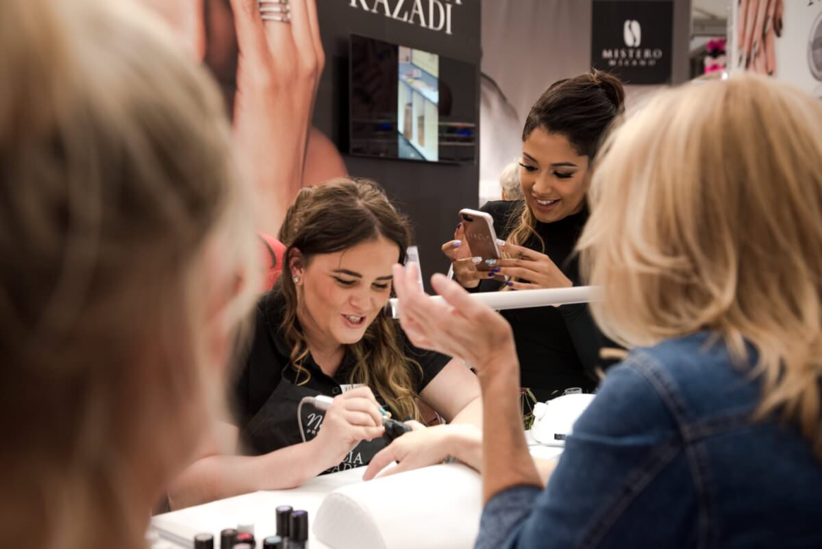 Kobieta robi sobie manicure w salonie, a obok stoi inna kobieta, fotograf eventowy, uśmiechając się i robiąc zdjęcie telefonem. W tle na ścianach widoczne są branding salonu i materiały promocyjne. Atmosfera jest spokojna i wesoła.