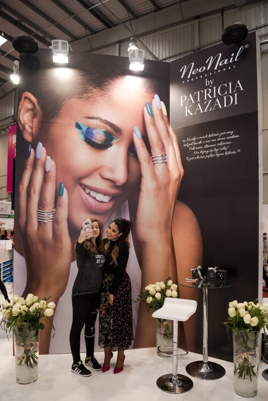 Dwie osoby stoją przed dużym stoiskiem NeoNail, na którym występuje Patricia Kazadi i utrwalają ten moment na selfie. Na wystawie widać uśmiechniętą modelkę z kolorowym makijażem oczu i wypielęgnowanymi paznokciami. Stoisko otoczone jest białymi różami, które pięknie uchwycił Marcin Krokowski, fotograf w Warszawie.